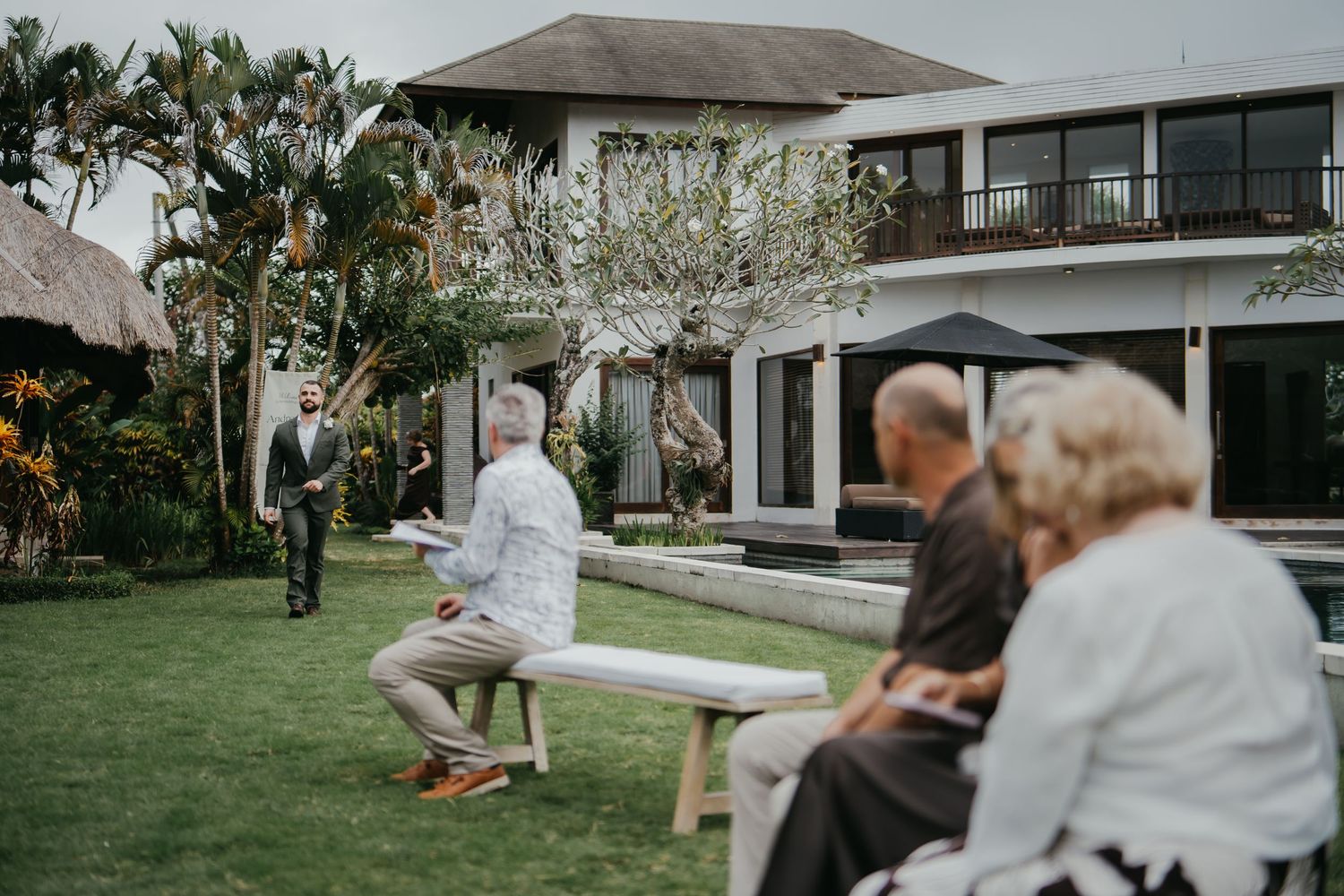 Wedding guests seated on benches as groom walks across lush garden toward ceremony.