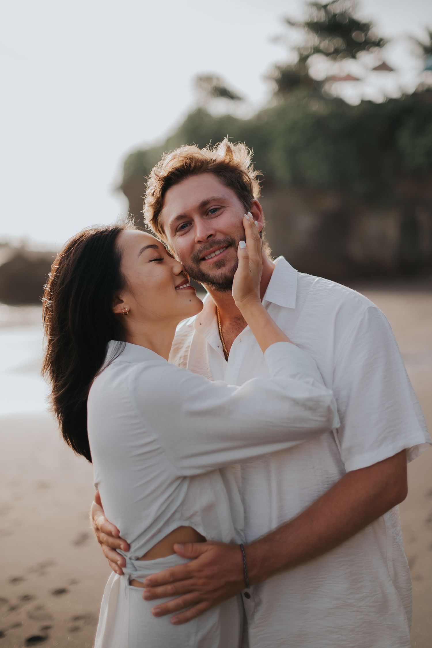 A romantic couple in white outfits share tender moments together on a sandy beach during sunset.