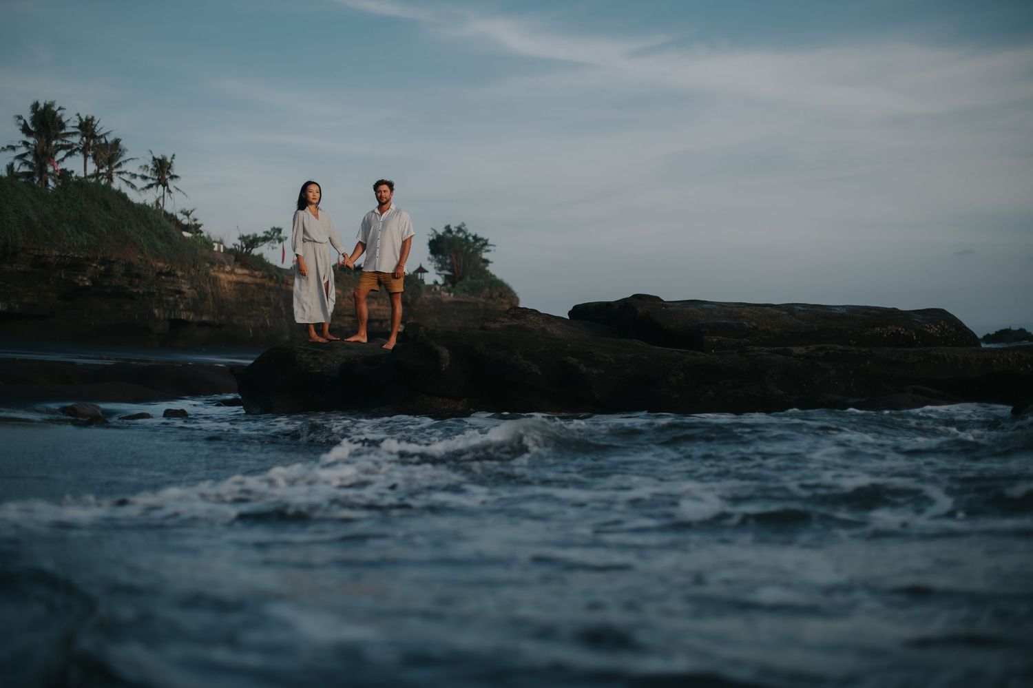 Couple sits on rocky ocean cliffs at sunset while waves crash against the shore in Hawaii.