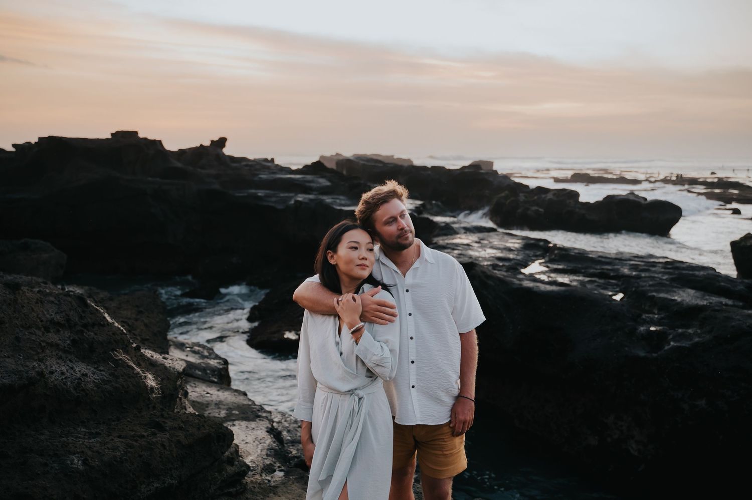 A couple shares an intimate moment on rocky coastal cliffs at sunset with ocean waves crashing in the background.