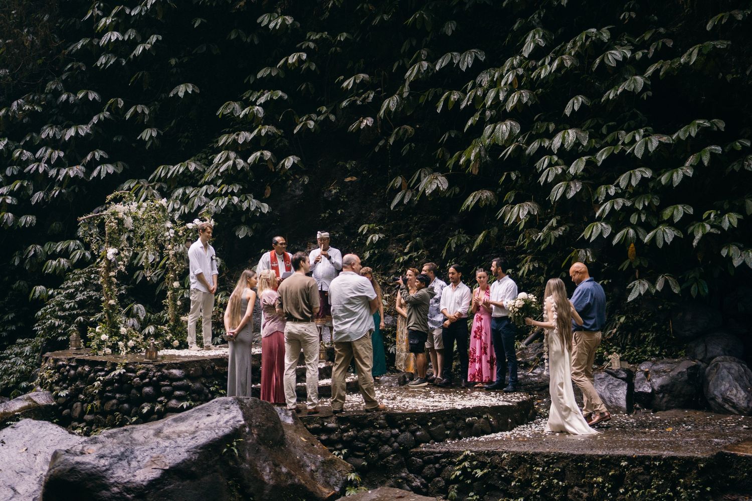 A wedding party stands in front of a lush green forest backdrop during an outdoor ceremony.