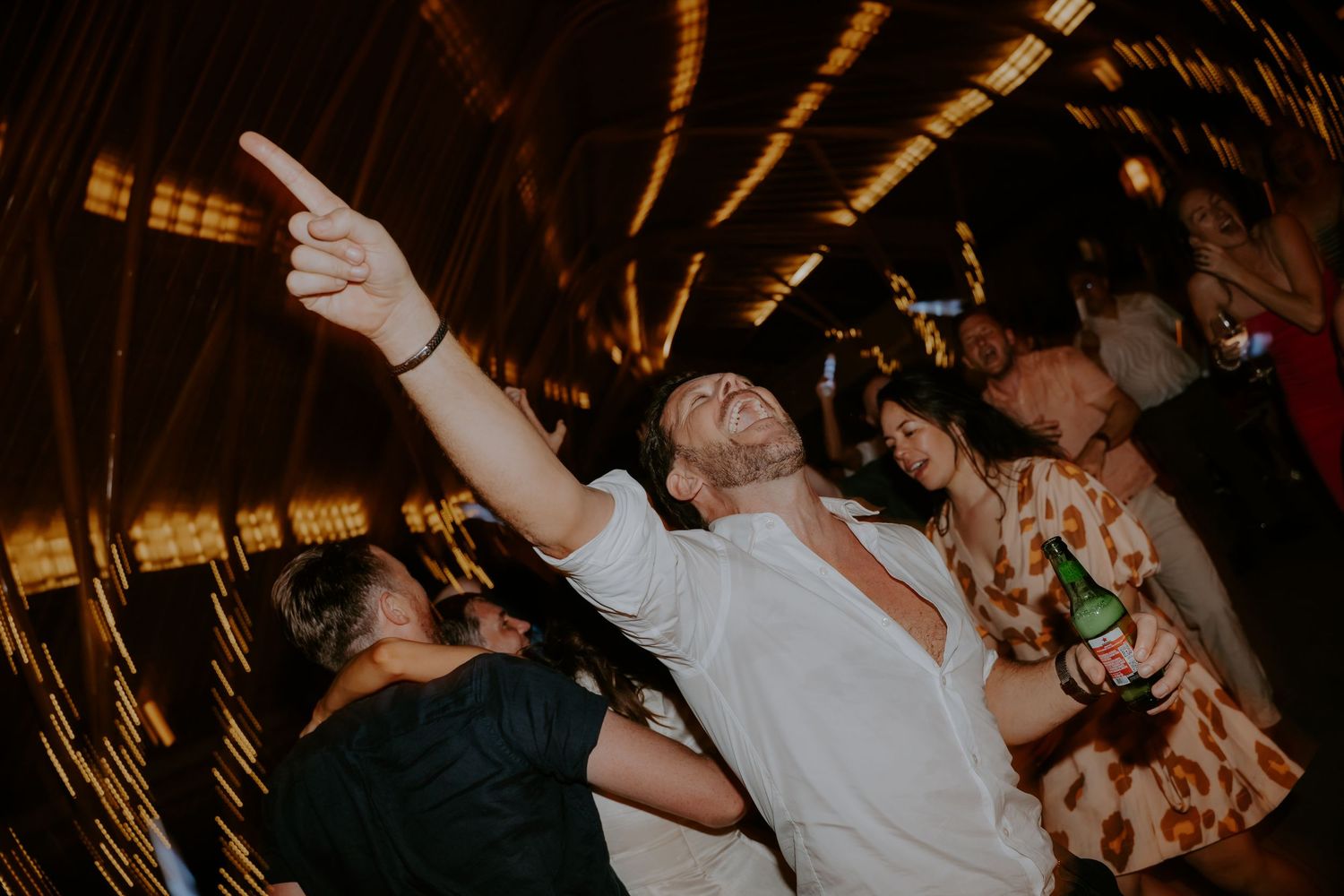 Energetic crowd dances with raised arms at a dimly lit nightclub or concert venue.