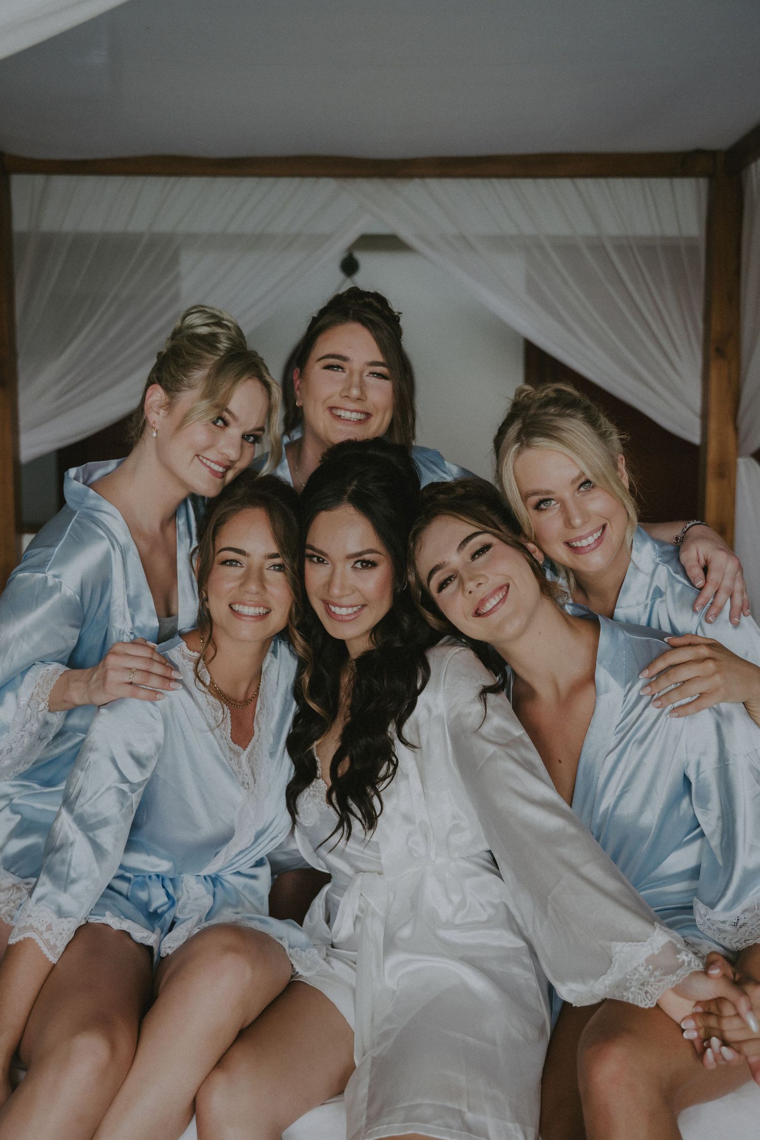 Group wearing matching silk robes pose together during wedding day preparations in light-filled room.