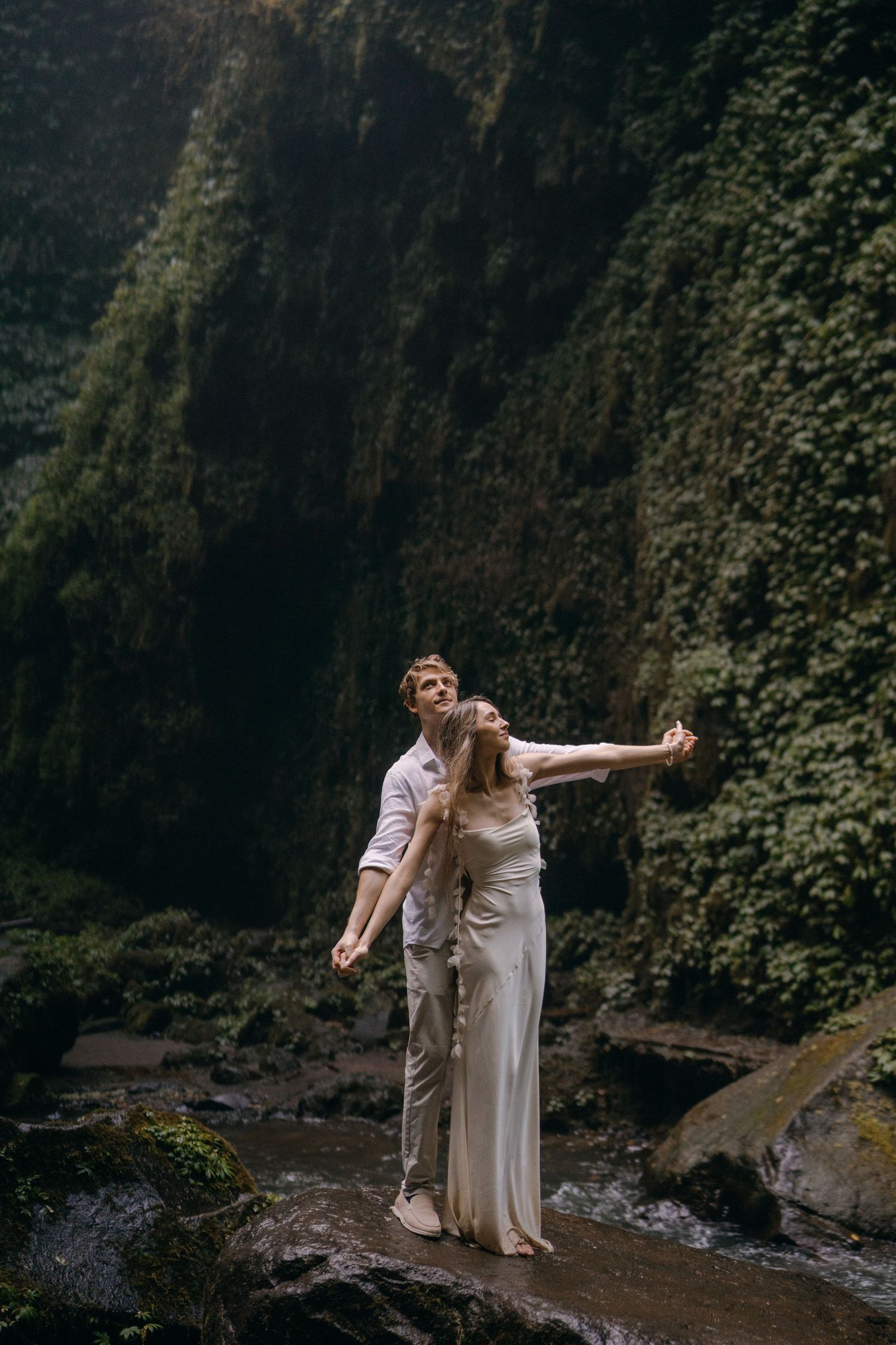 Couple in white outfits dancing and embracing by a scenic waterfall in a lush forest setting.