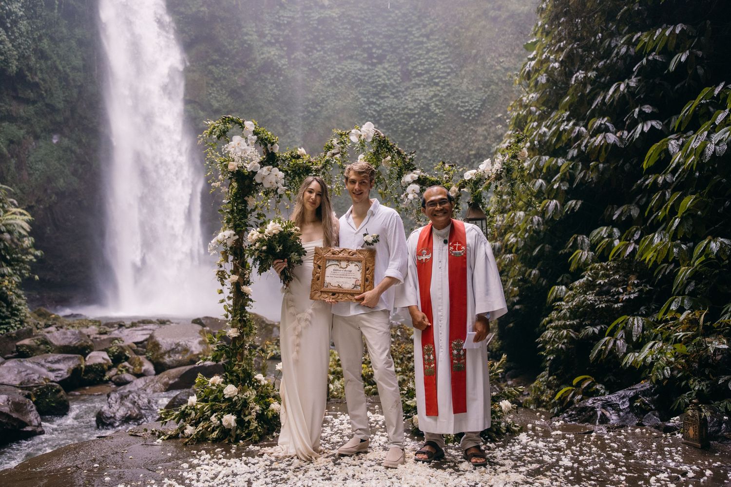 A couple stands in front of a majestic waterfall during their outdoor wedding ceremony in a lush natural setting.