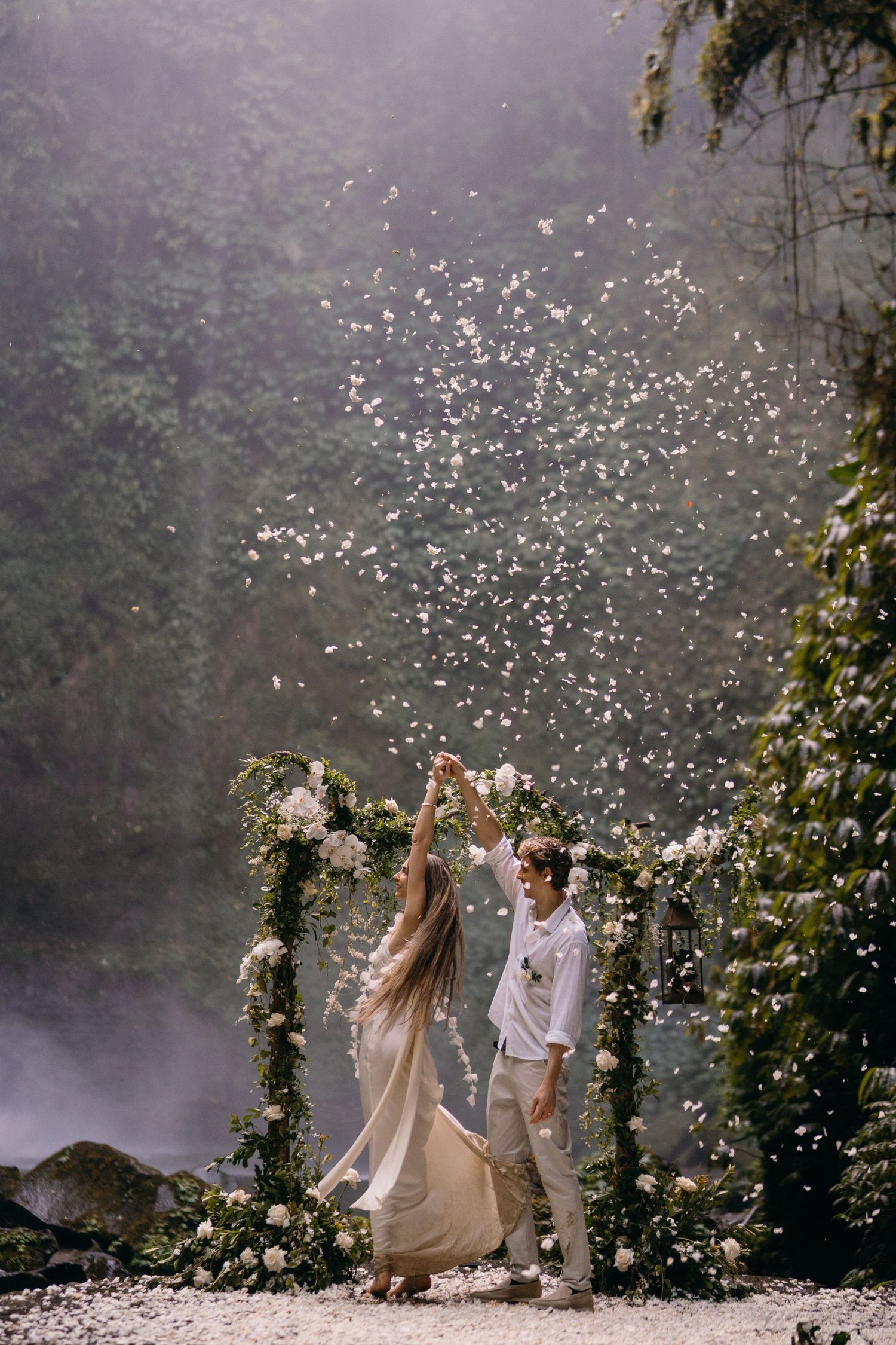 A couple celebrates under a floral arch by a misty waterfall as flower petals float through the ethereal scene.