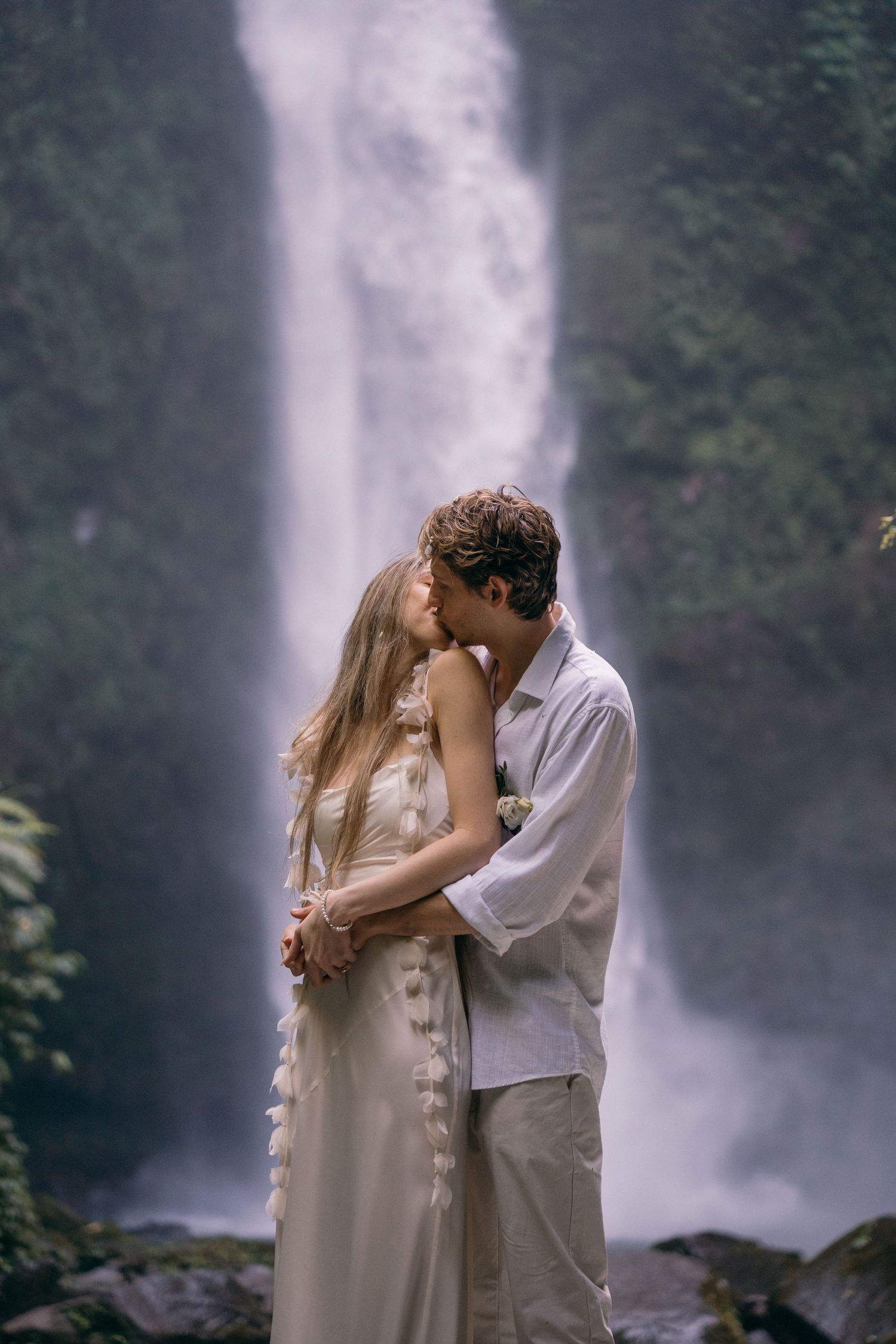 A couple shares a romantic kiss against the backdrop of a misty waterfall in a lush forest setting.