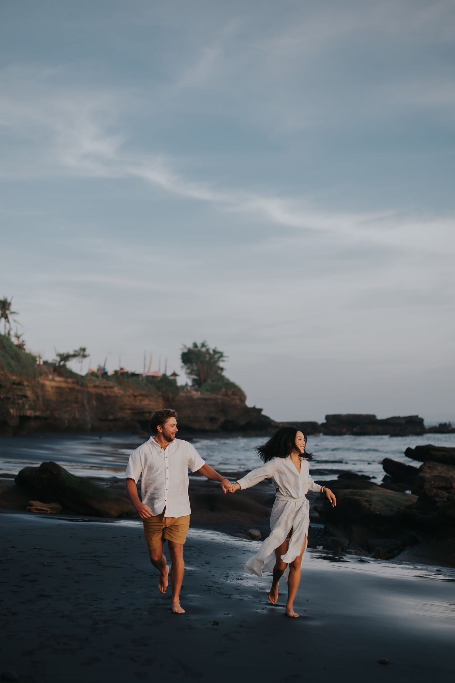 A couple playfully runs and dances together on a black sand beach with rocky cliffs and ocean in the background.