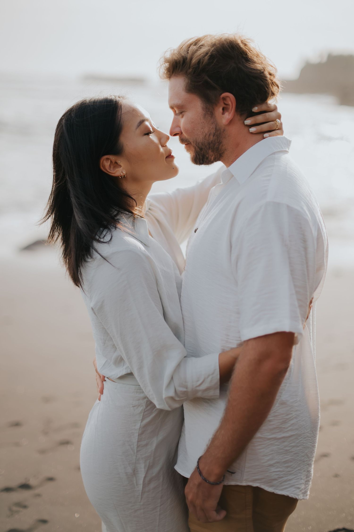 A romantic couple in white outfits share intimate moments on a misty beach at sunset.