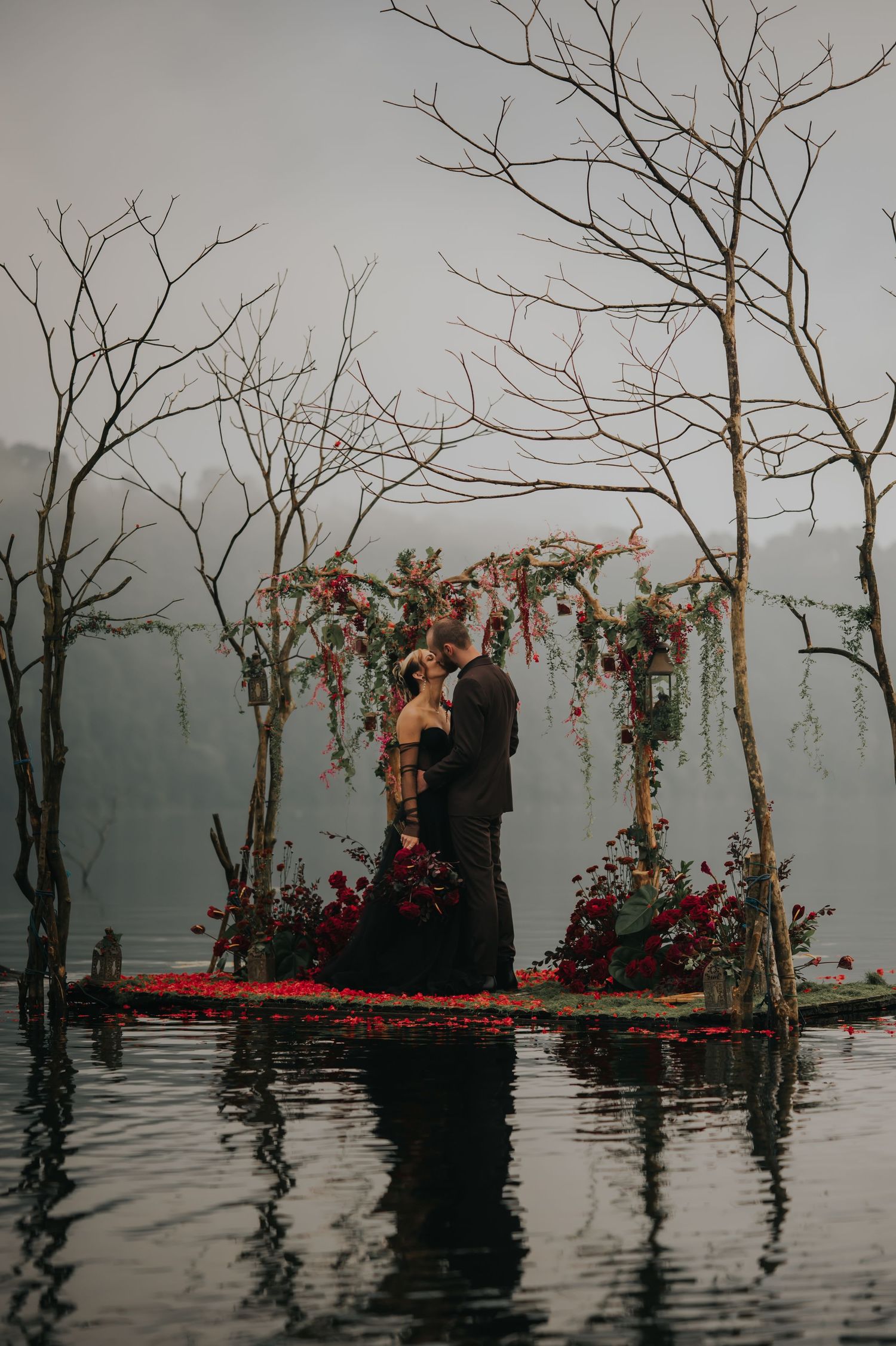 Moody wedding ceremony on a floating platform over misty water with bare trees and floral arch decorations.