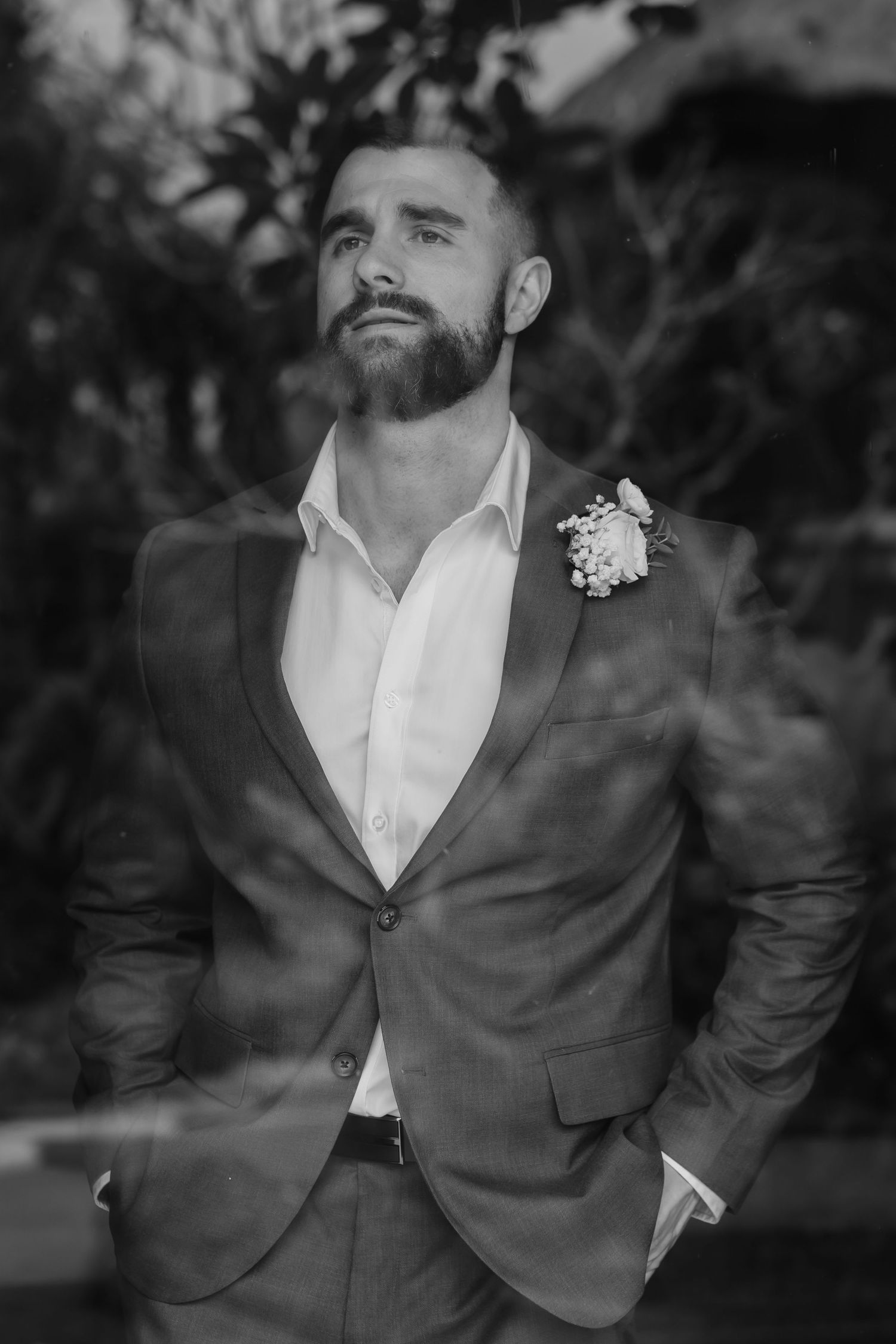 Bearded groom in grey suit with boutonniere poses outdoors in black and white wedding portrait.