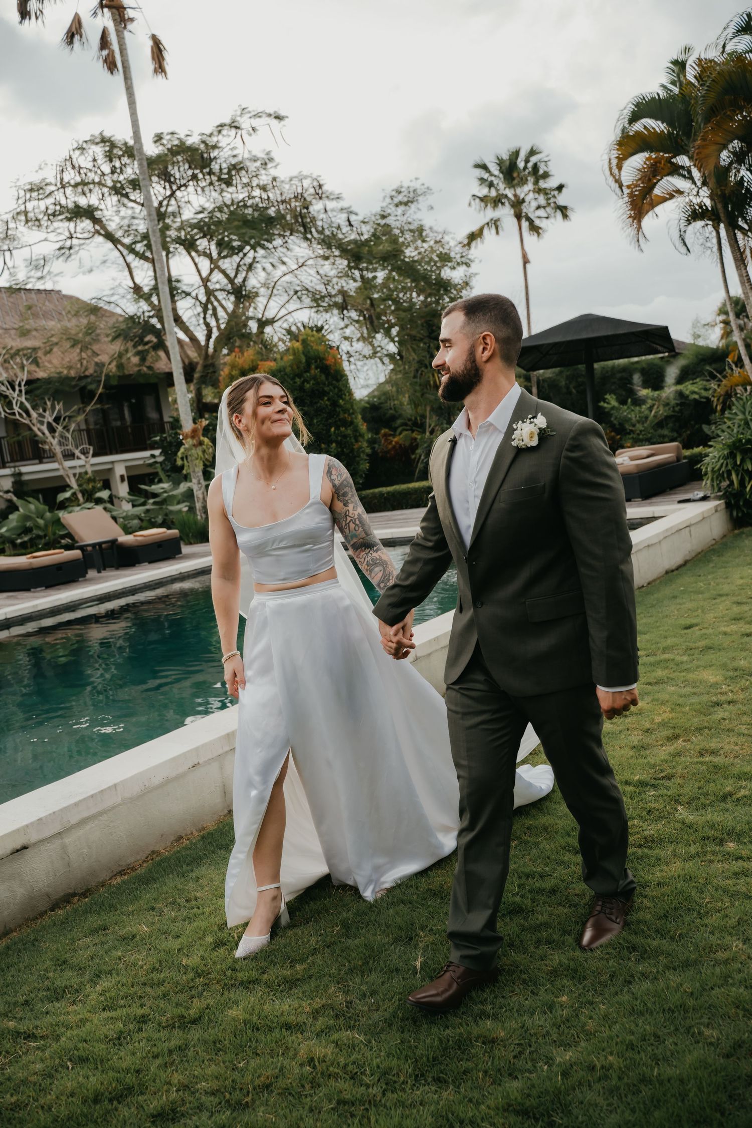 Bride and groom walk hand-in-hand by a pool, bride in a two-piece white gown, groom in a dark suit.