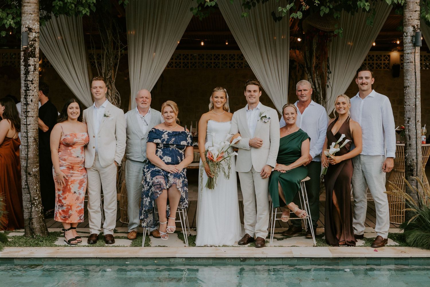 Bride and groom pose with family group photo beside pool at tropical outdoor wedding venue.