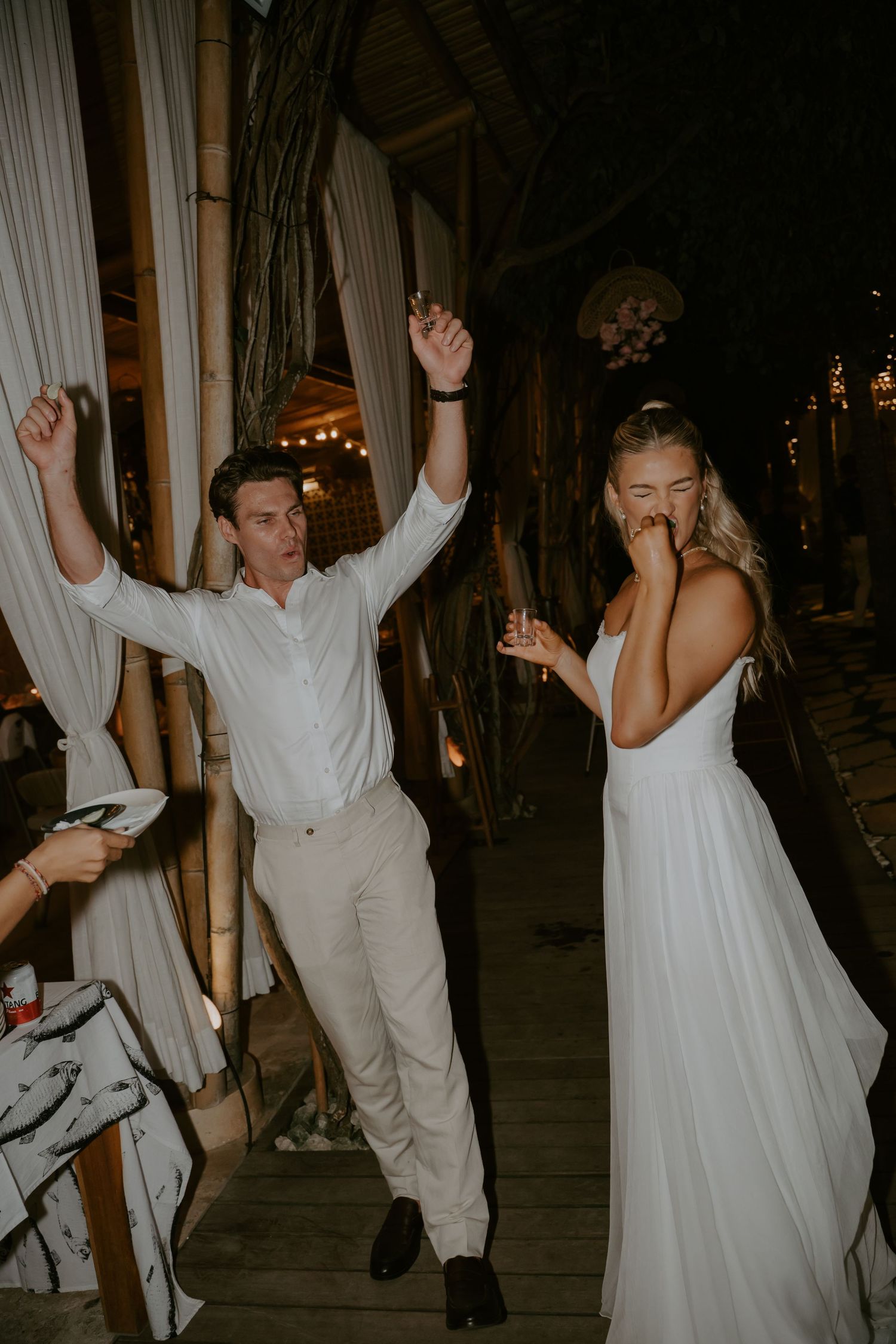 Joyful groom raises arms in celebration as laughing bride in white gown makes grand entrance at outdoor wedding reception.