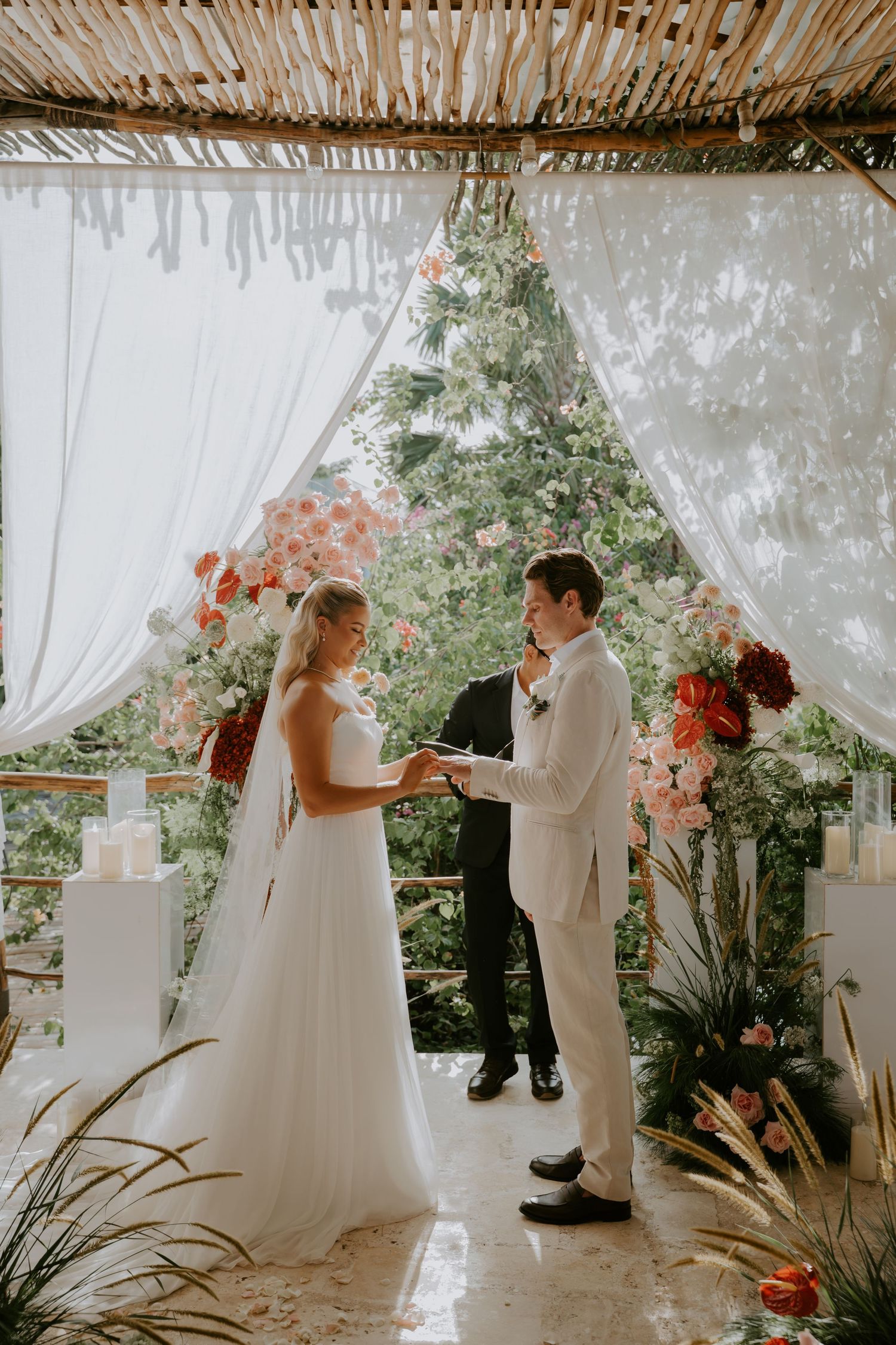 Bride and groom exchange rings under white draped altar adorned with pink and red floral arrangements.