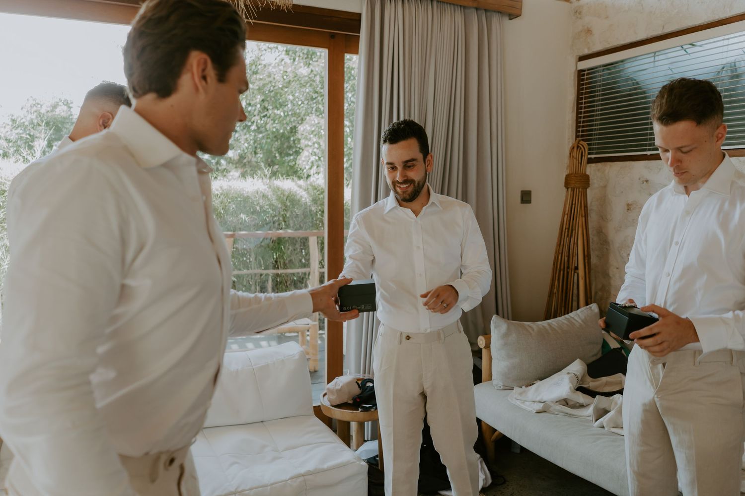 Groomsmen in white shirts and trousers prepare together in a bright villa room before the wedding ceremony.