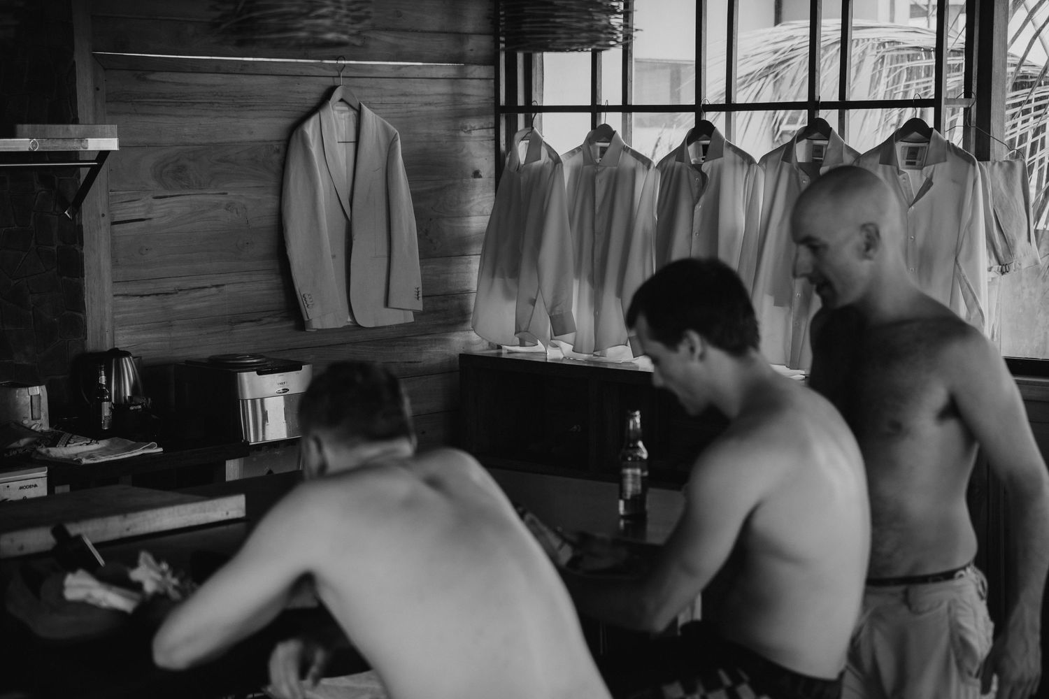 Three shirtless men in a rustic wooden room with suits hanging in the background, black and white photo.