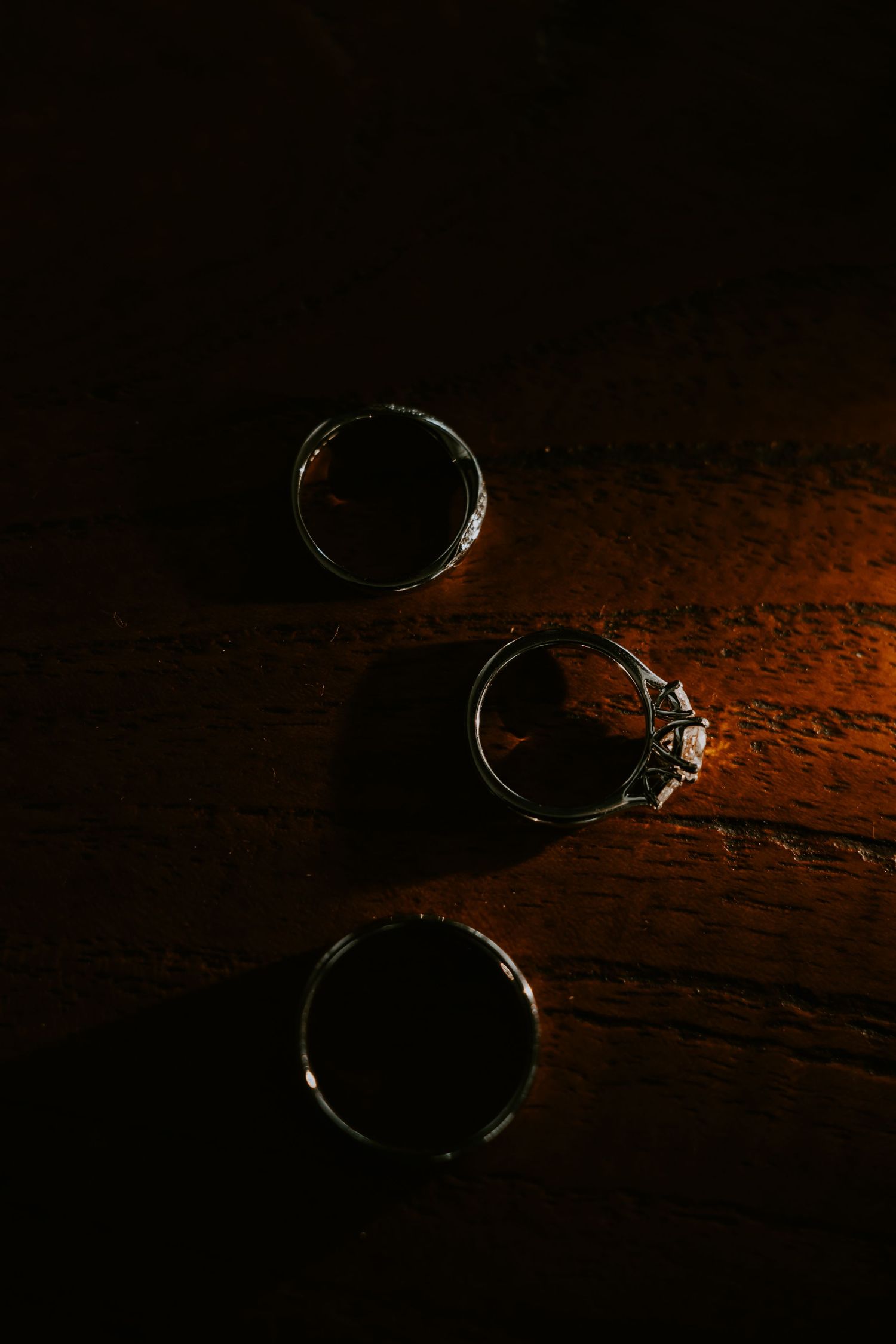 Two wedding rings and an engagement ring with diamond displayed on dark wooden surface in dramatic lighting.