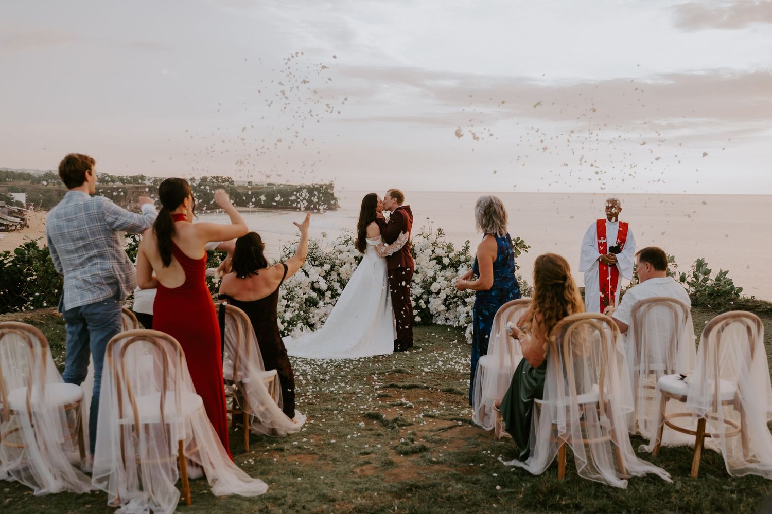 Wedding guests toss confetti as couple shares first kiss at stunning cliffside outdoor ceremony at sunset.