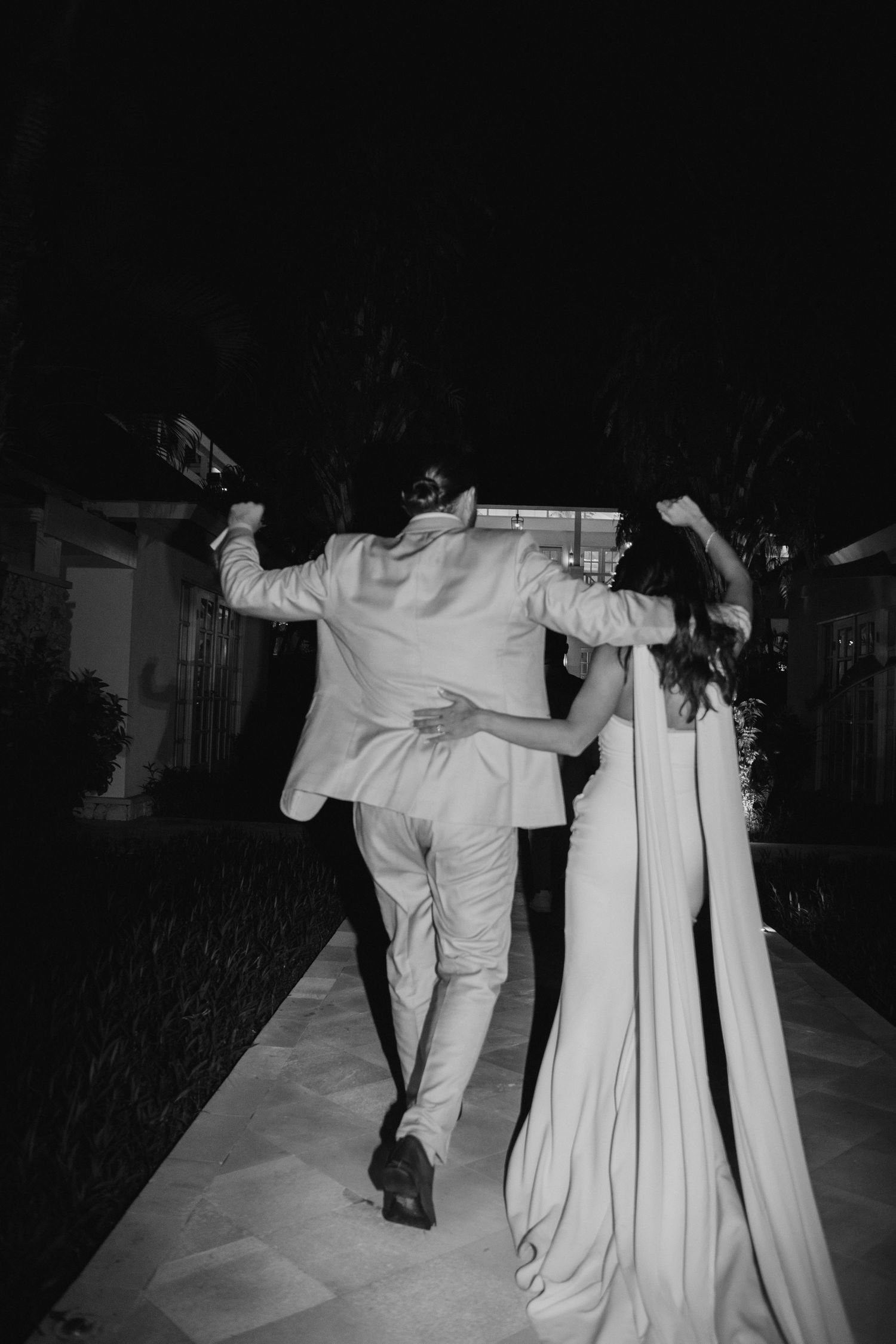 Black and white photo of a newlywed couple walking joyfully together along a path at night.