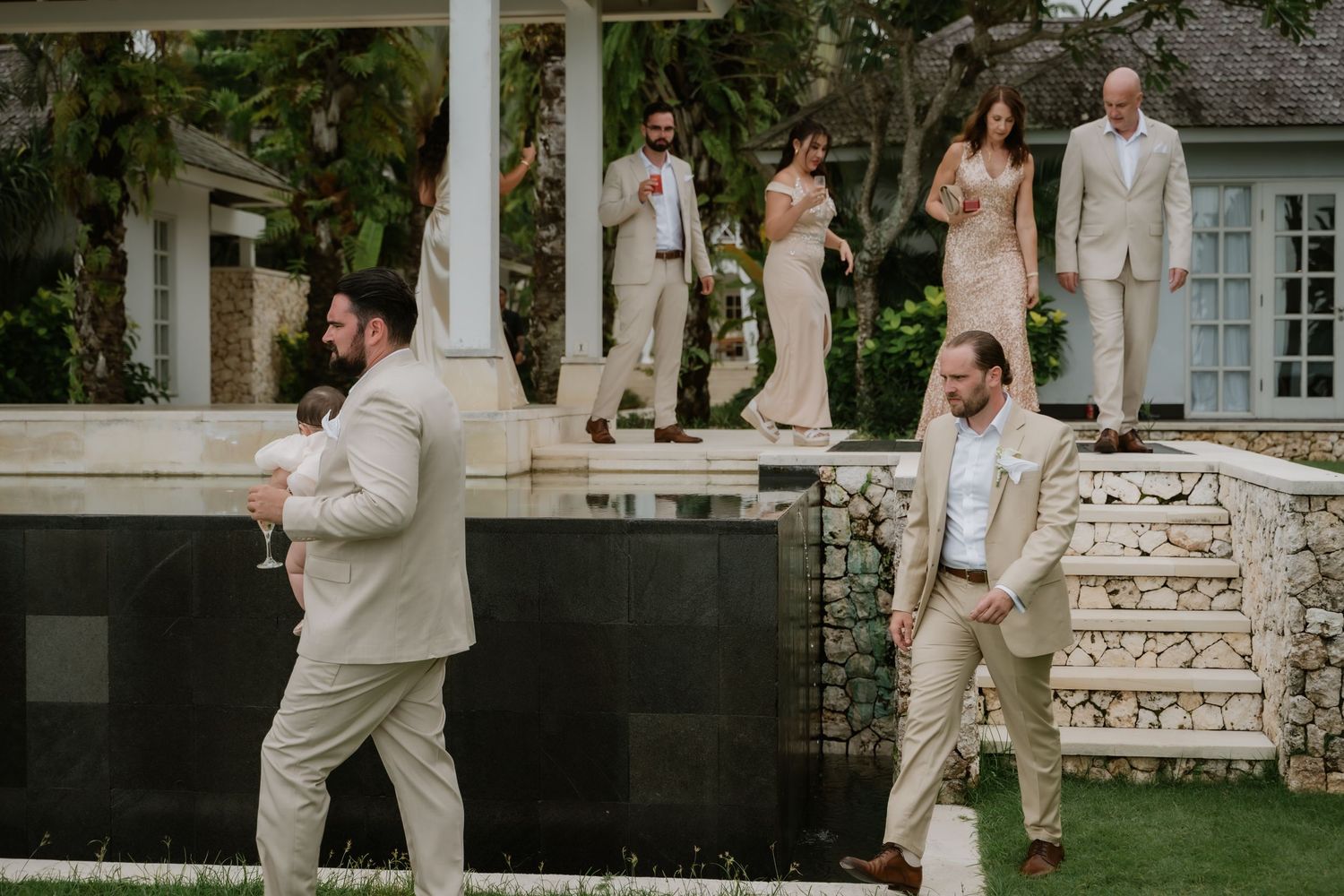 Wedding party members in tan suits and gold dresses gather at tropical venue.