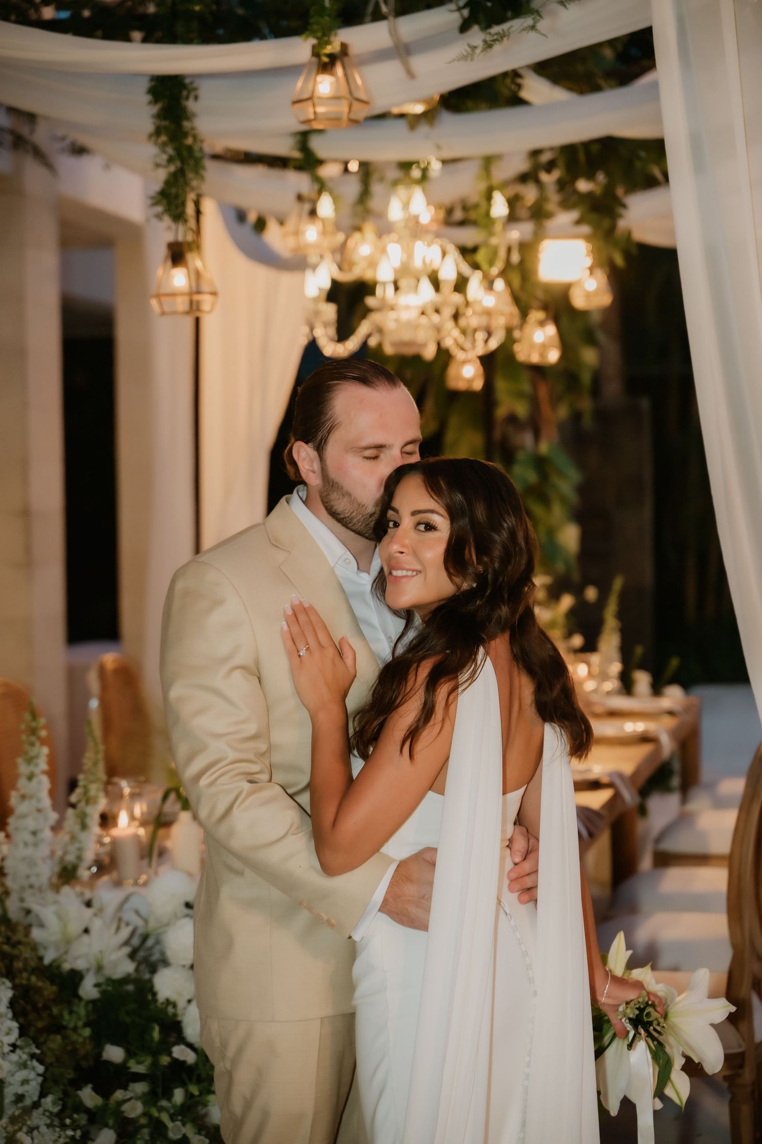 Groom in beige suit embraces smiling bride under chandelier lights at romantic evening reception.