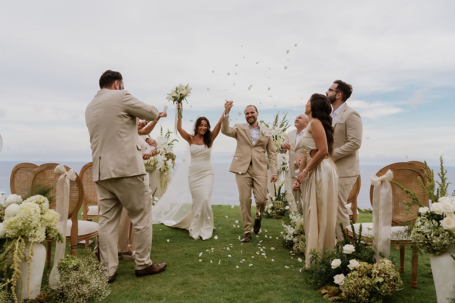 Joyful couple celebrates wedding ceremony exit with raised hands while guests toss petals outdoors.