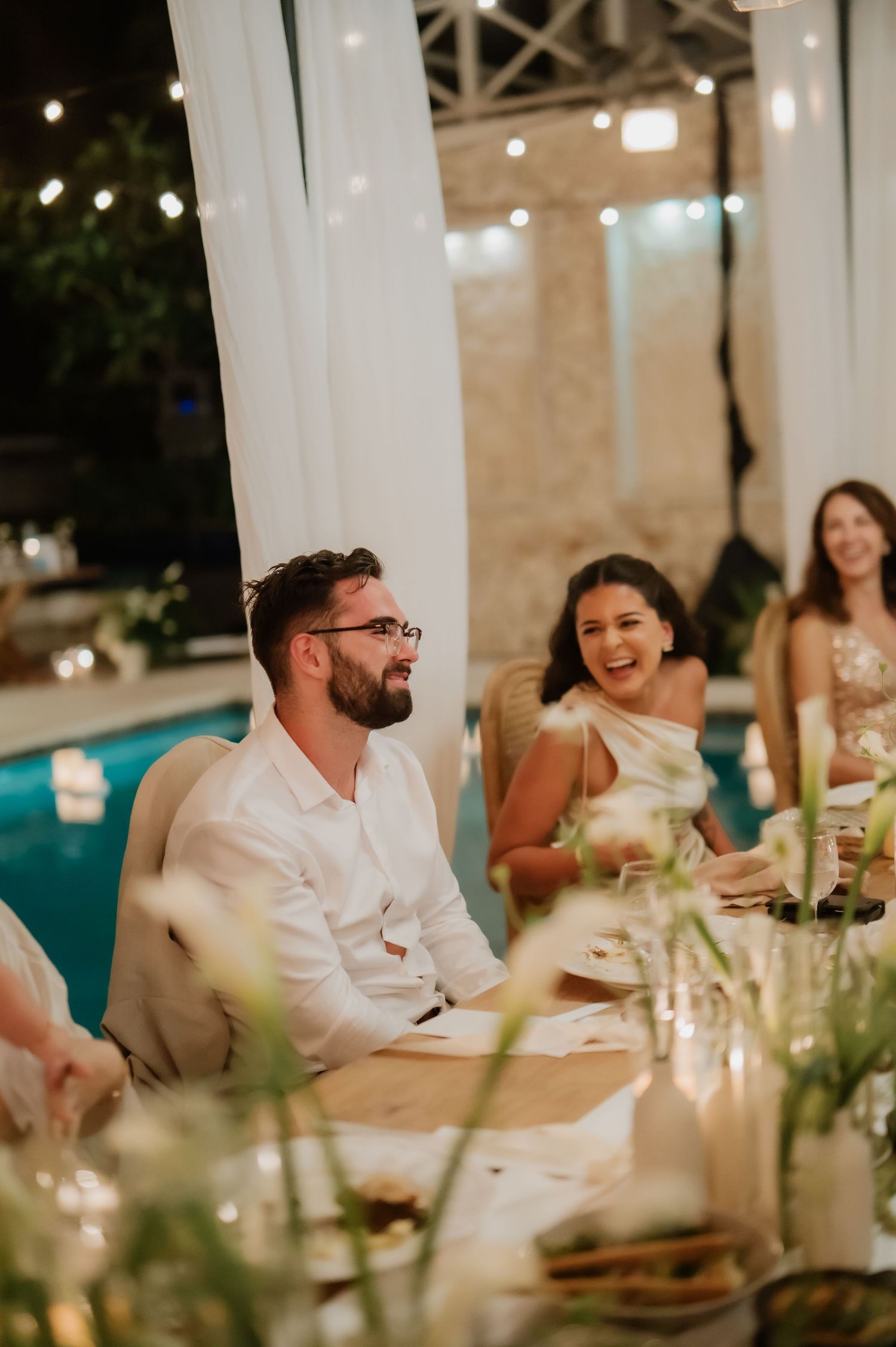 Wedding guests laugh joyfully during poolside evening reception with string lights and elegant table.