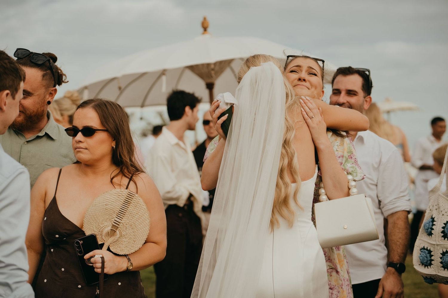 Bride in white veil embraces guest outdoors at wedding reception with umbrella tent in background.
