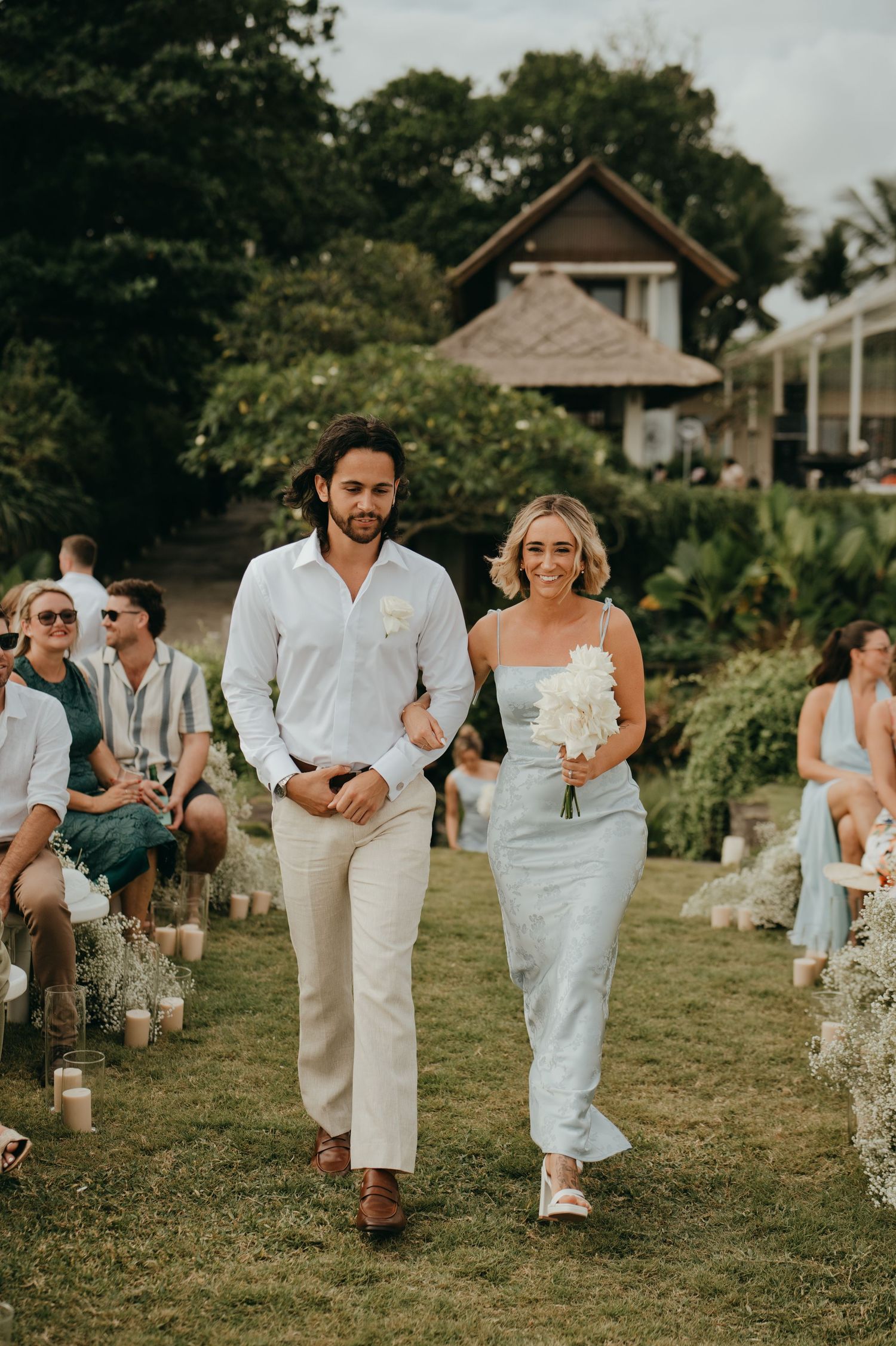 Bride in blue dress and groom in white shirt walk down grass aisle at tropical outdoor wedding ceremony.