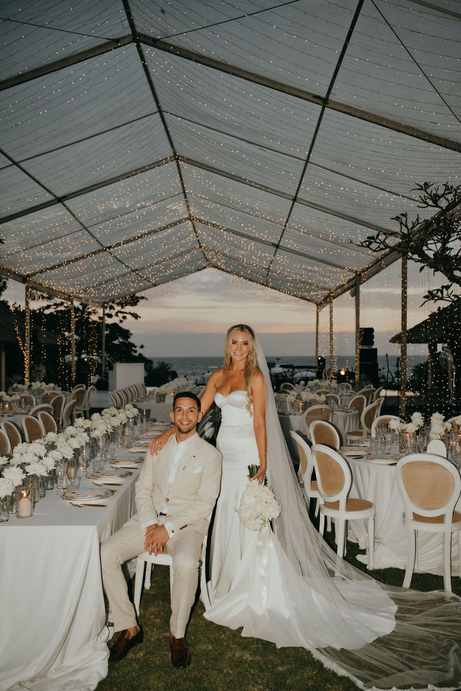 Bride and groom posing under fairy light canopy at twilight outdoor wedding reception.