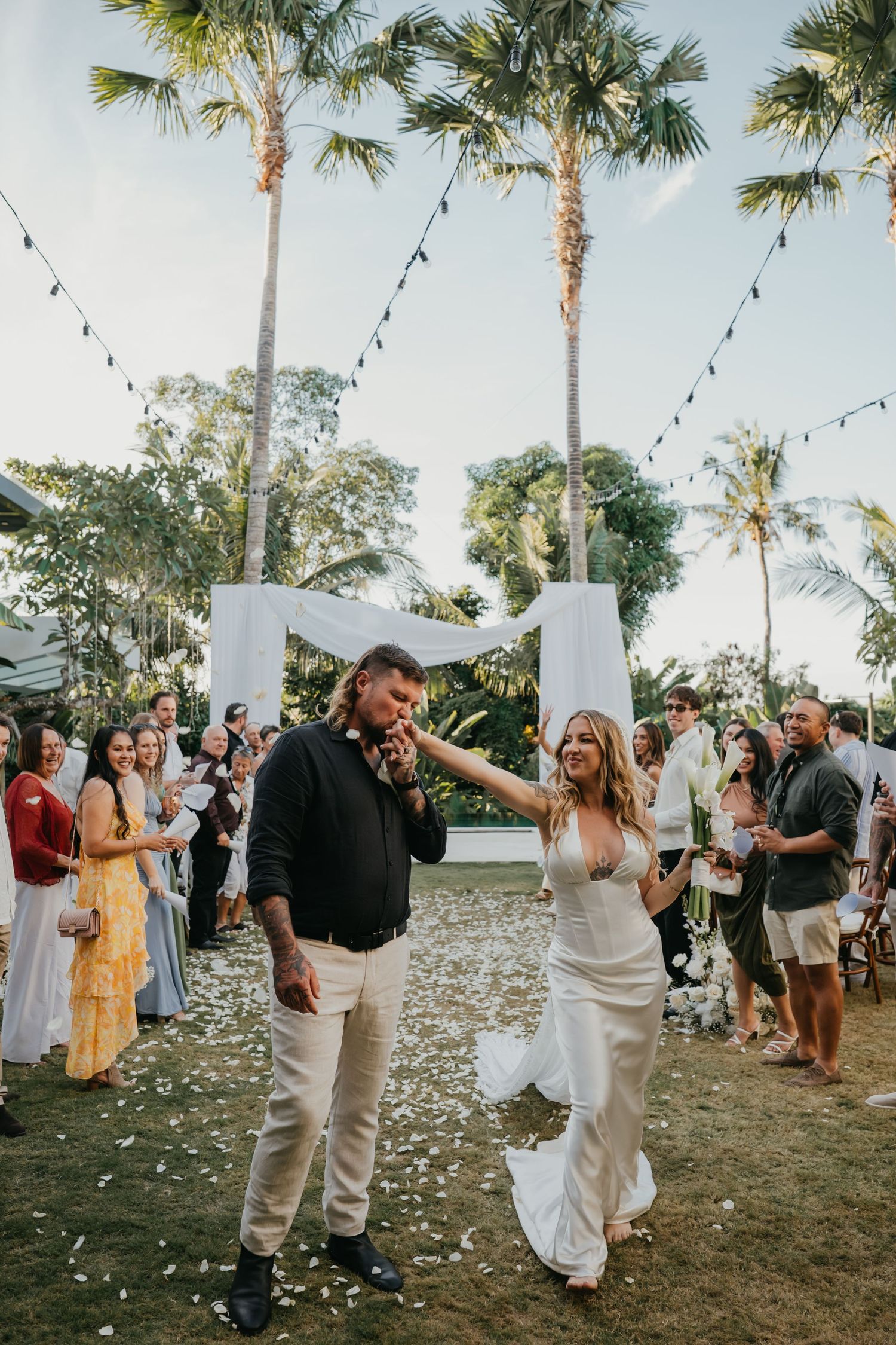Newlyweds walk down aisle amid cheering guests at tropical outdoor wedding ceremony with palm trees.