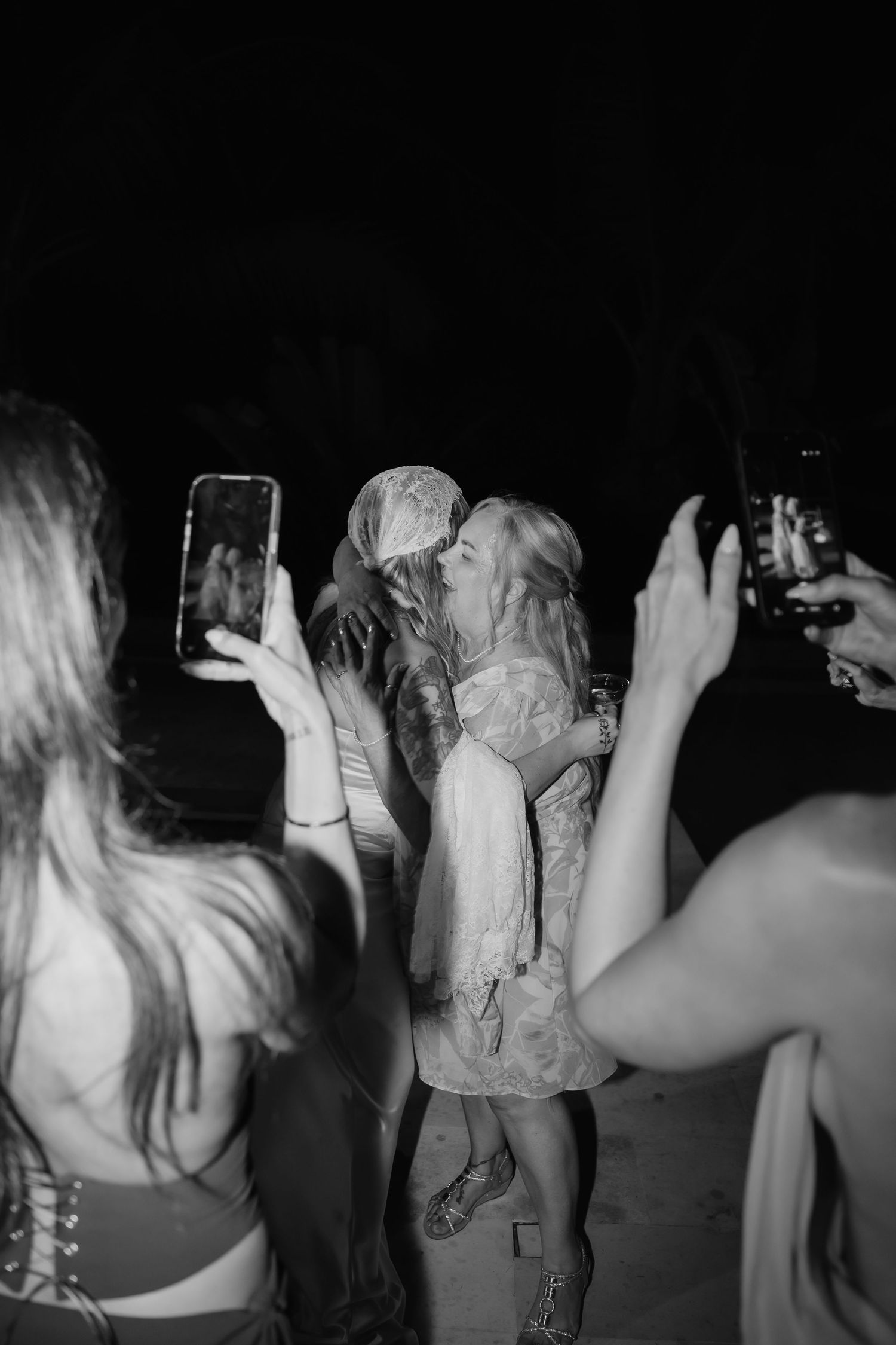 Black and white photo of newlyweds kissing while guests photograph the moment at night.
