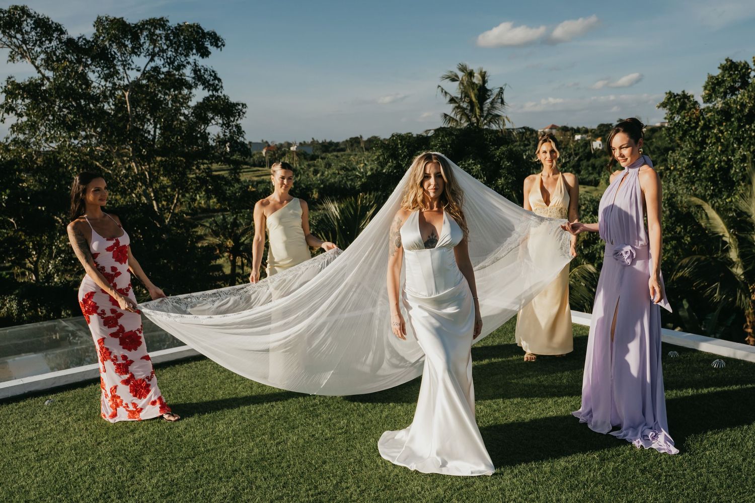 Bride in fitted white gown and veil with bridesmaids in colorful dresses on lush green lawn.