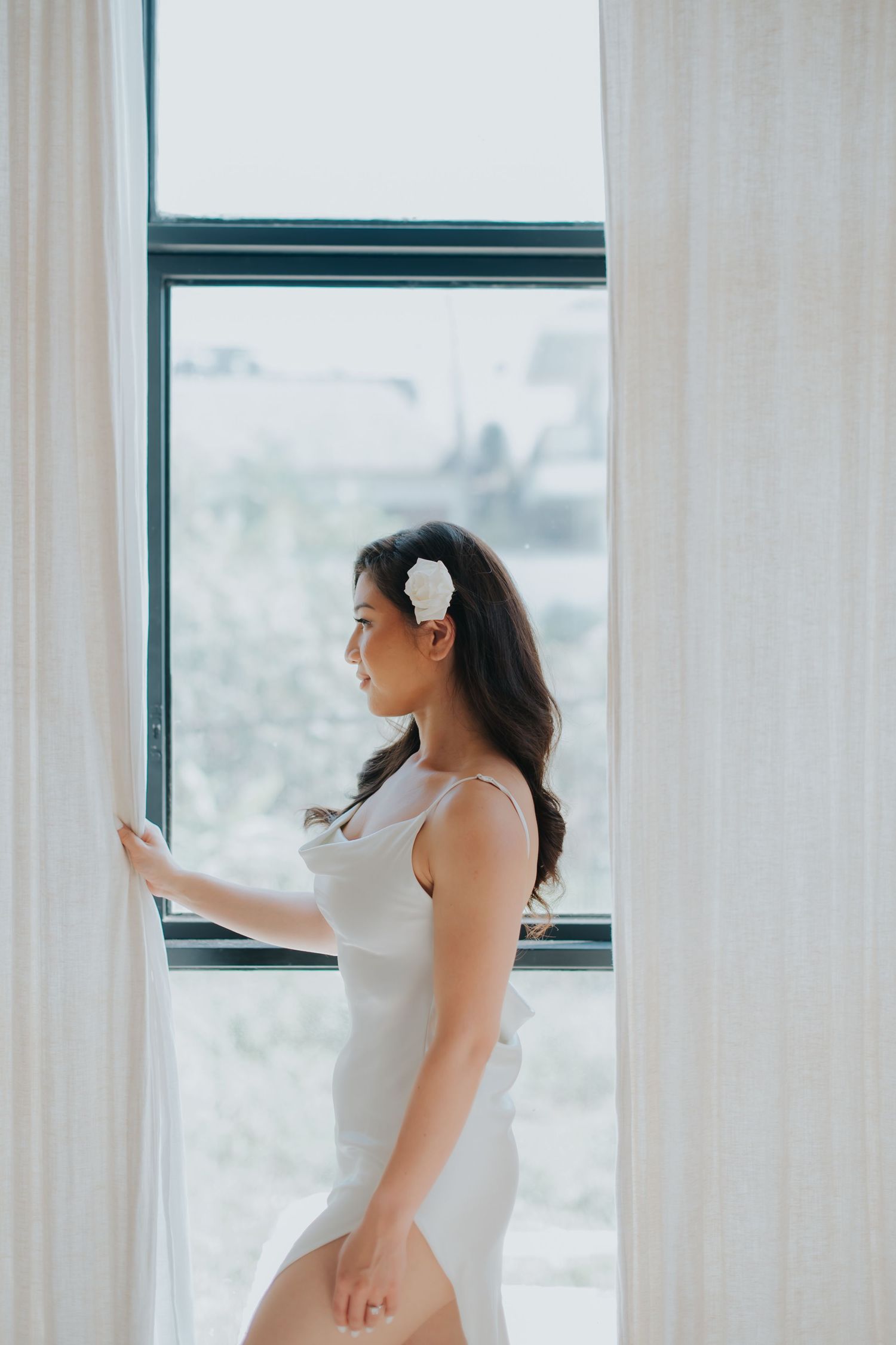 A woman in a white dress with a floral hair accessory stands by a bright window, holding sheer curtains.