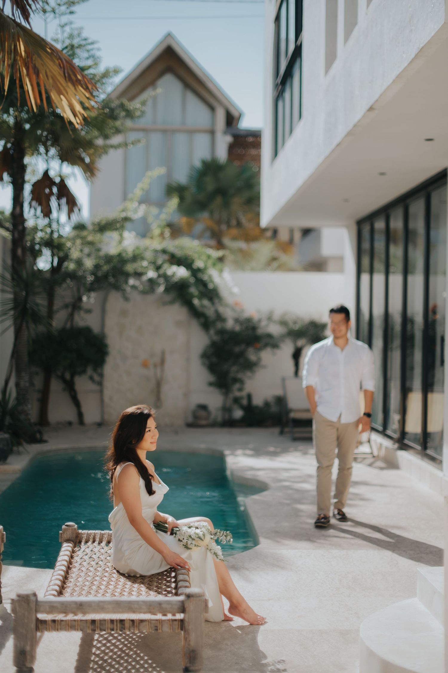 A woman sits on a wooden lounger by a pool while a man walks toward her in a modern tropical villa courtyard.