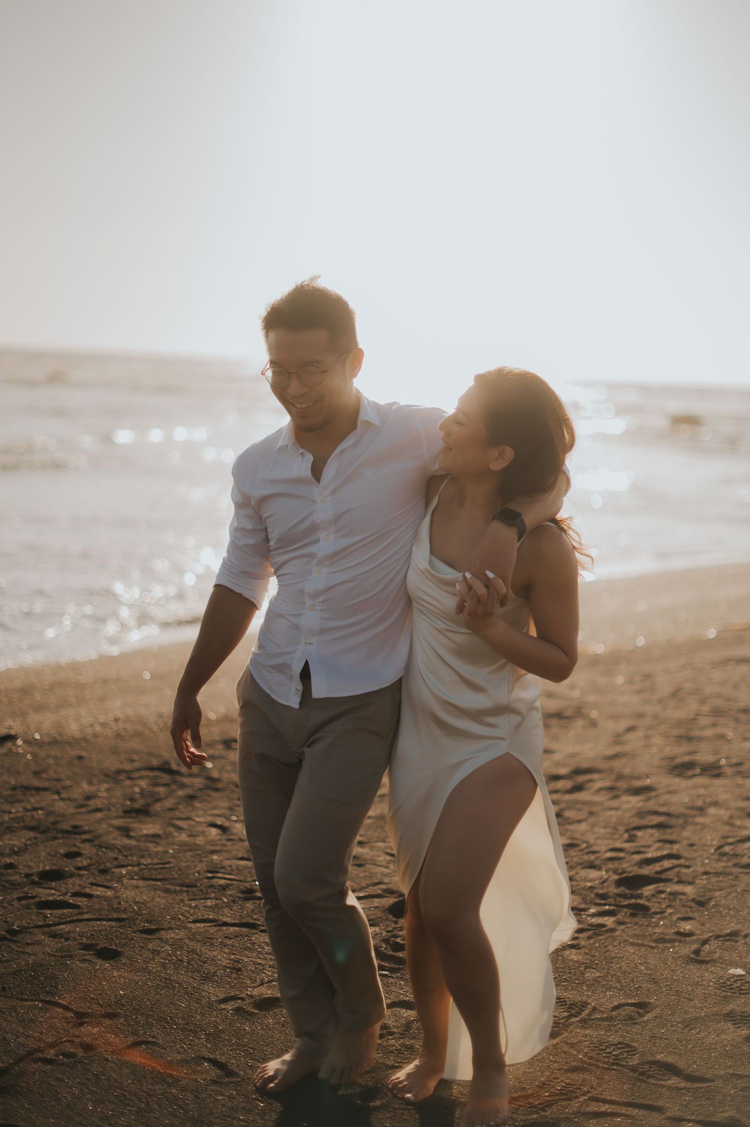 A couple walks barefoot on a sandy beach at sunset, the woman in a white dress embracing the smiling man in a white shirt.
