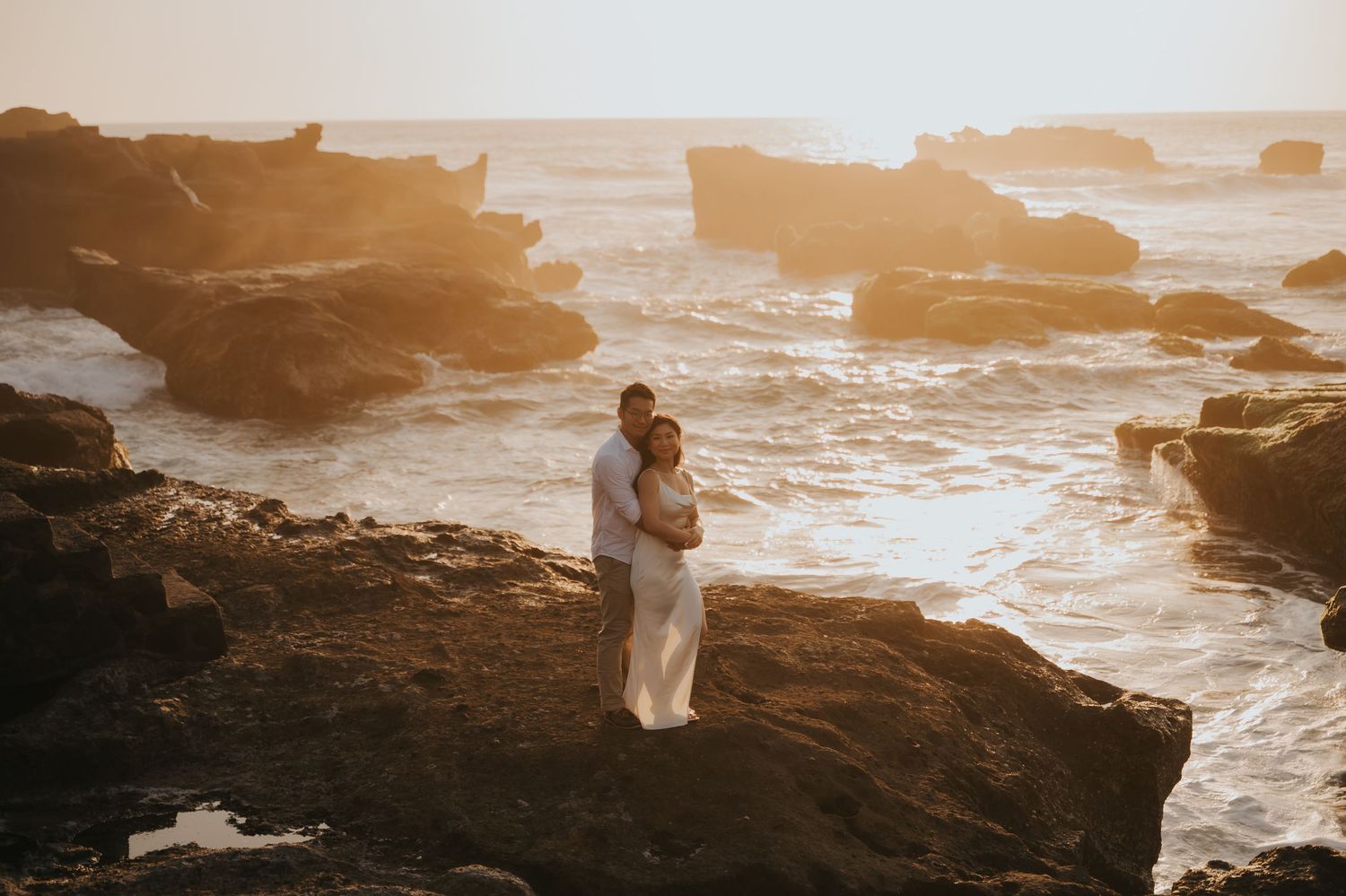 Couple embracing on rocky coastal cliffs at golden sunset with dramatic ocean waves crashing behind them.