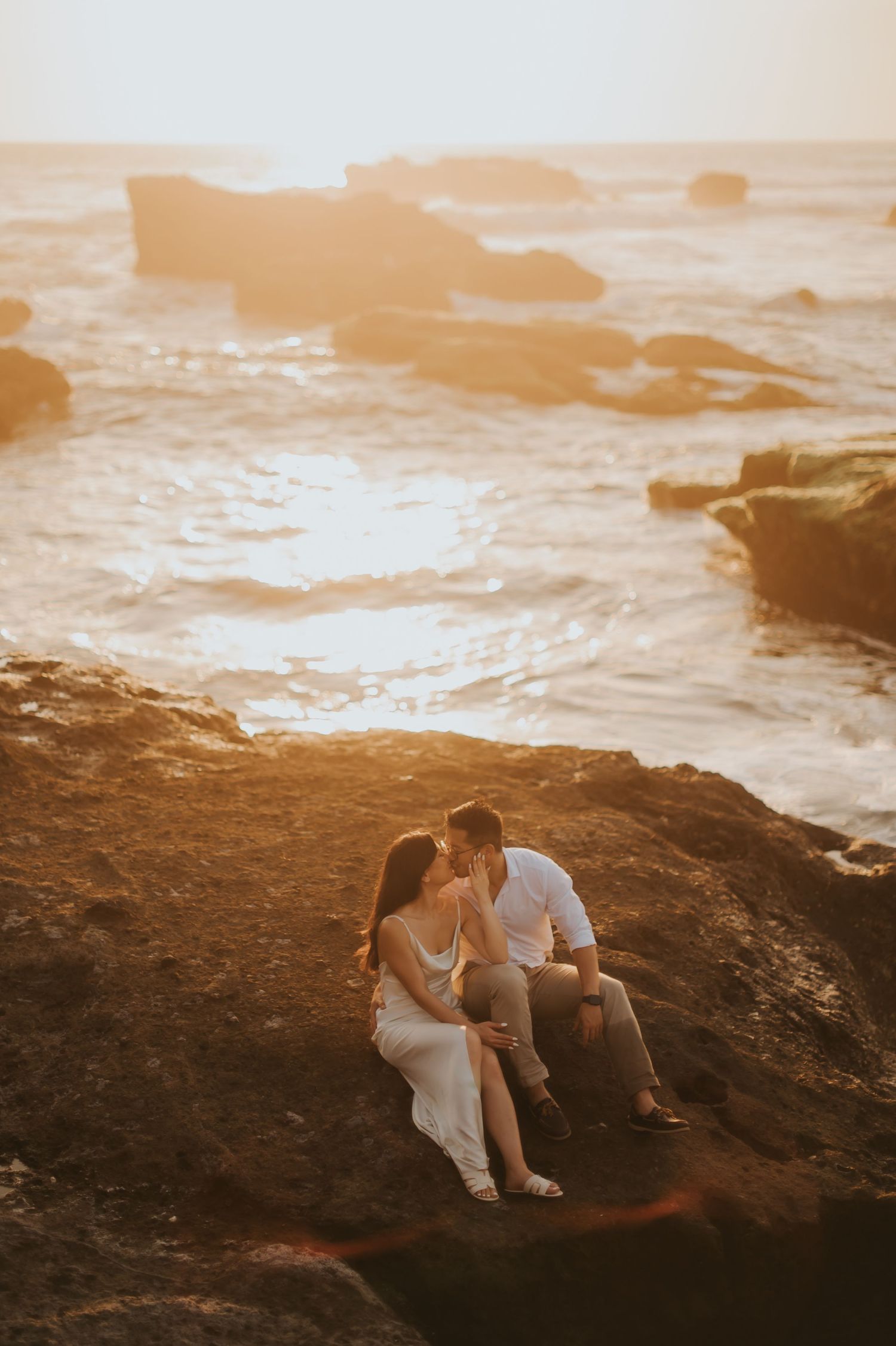 Couple sharing a romantic moment on rocky coastal cliffs at golden sunset with ocean waves in background.
