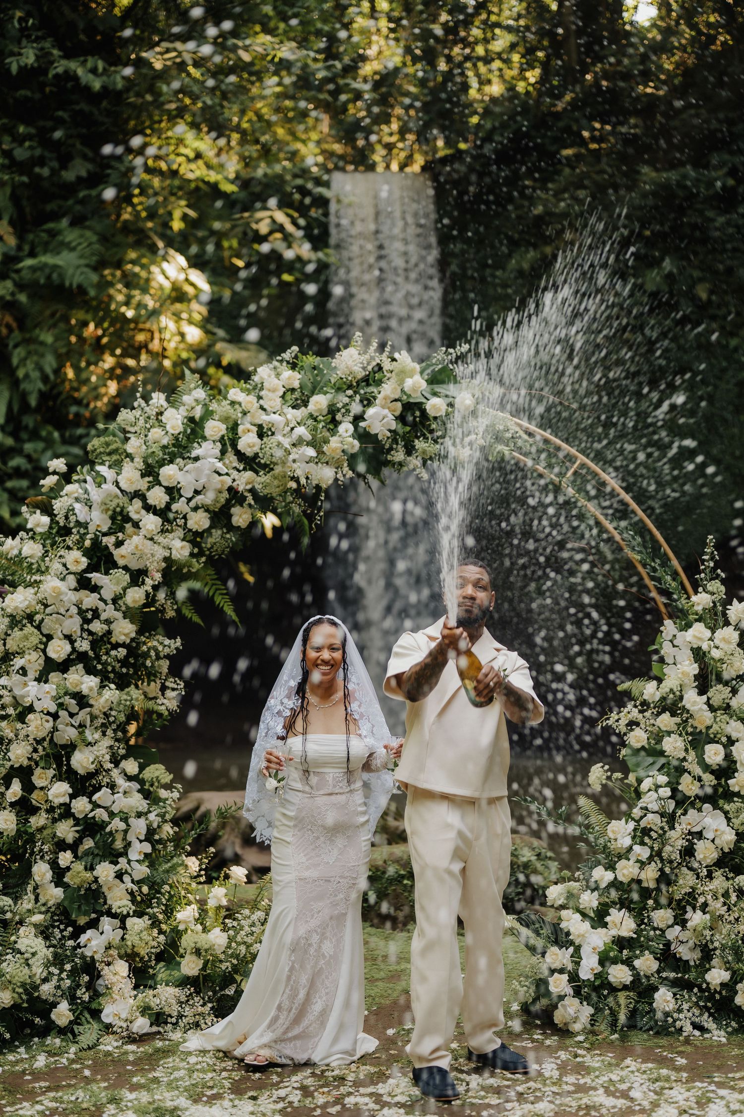 Bride and groom celebrating with champagne spray at outdoor wedding altar adorned with white flowers and waterfall backdrop.