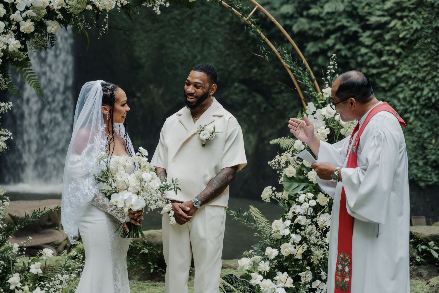 Bride and groom exchange vows before priest at elegant outdoor wedding with circular floral arch.
