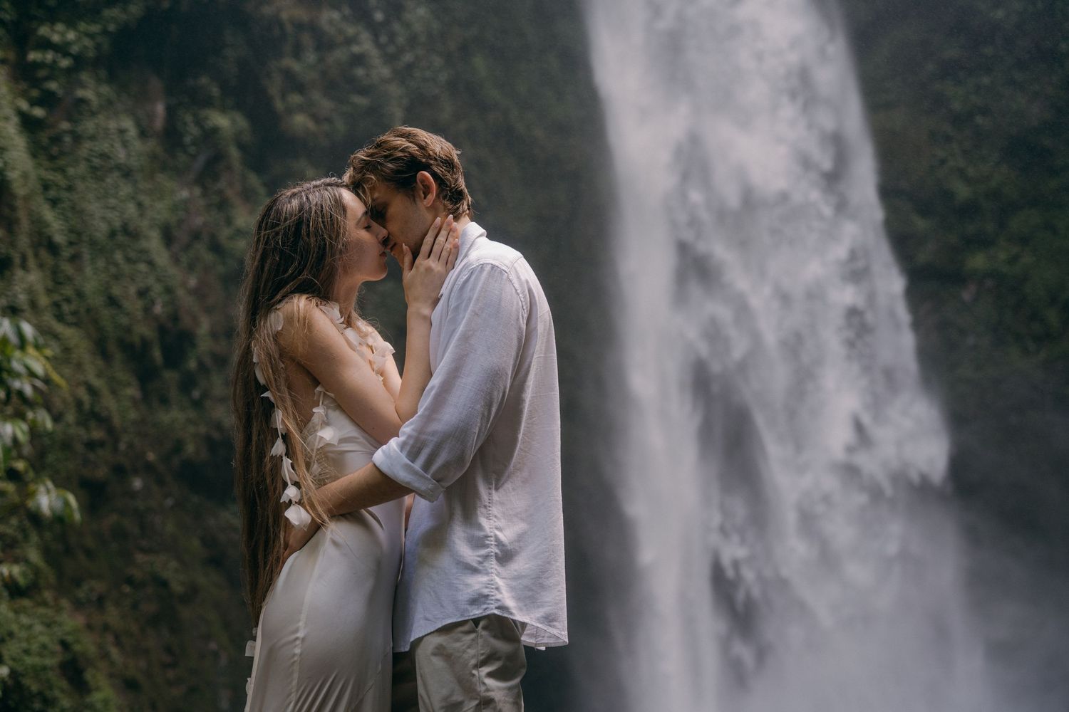 A romantic couple embraces in front of a majestic waterfall with mist rising in the background.