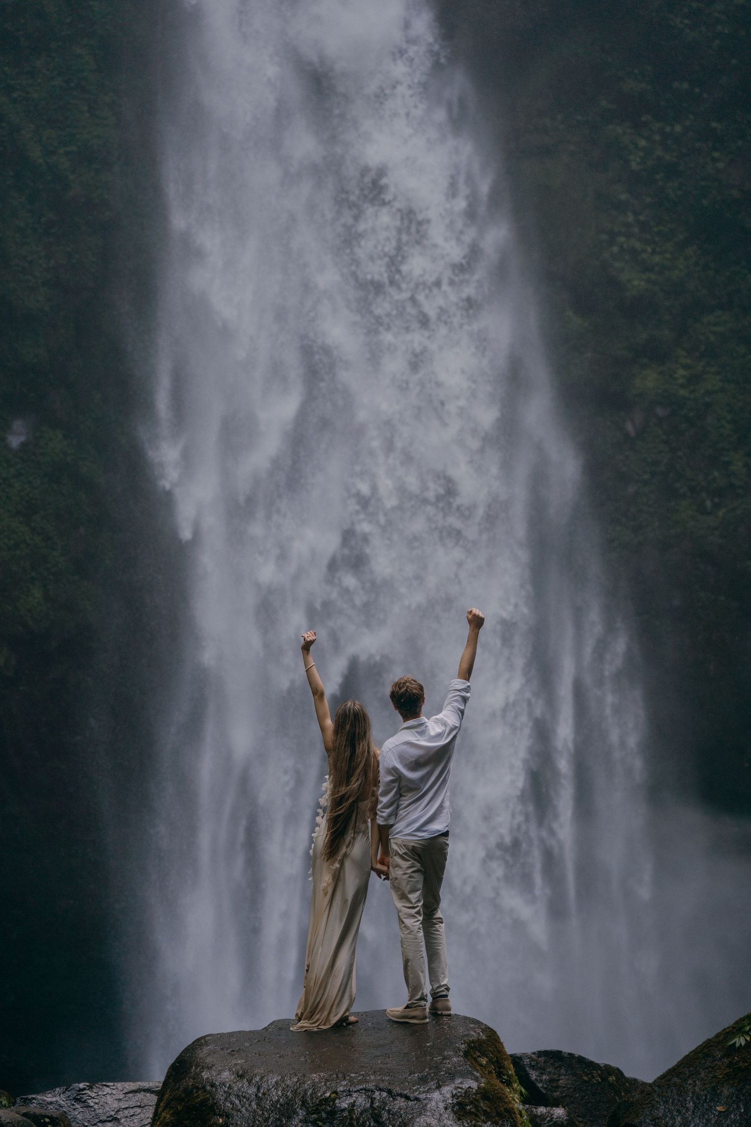 Two people stand triumphantly with raised arms in front of a majestic misty waterfall in a dramatic nature scene.