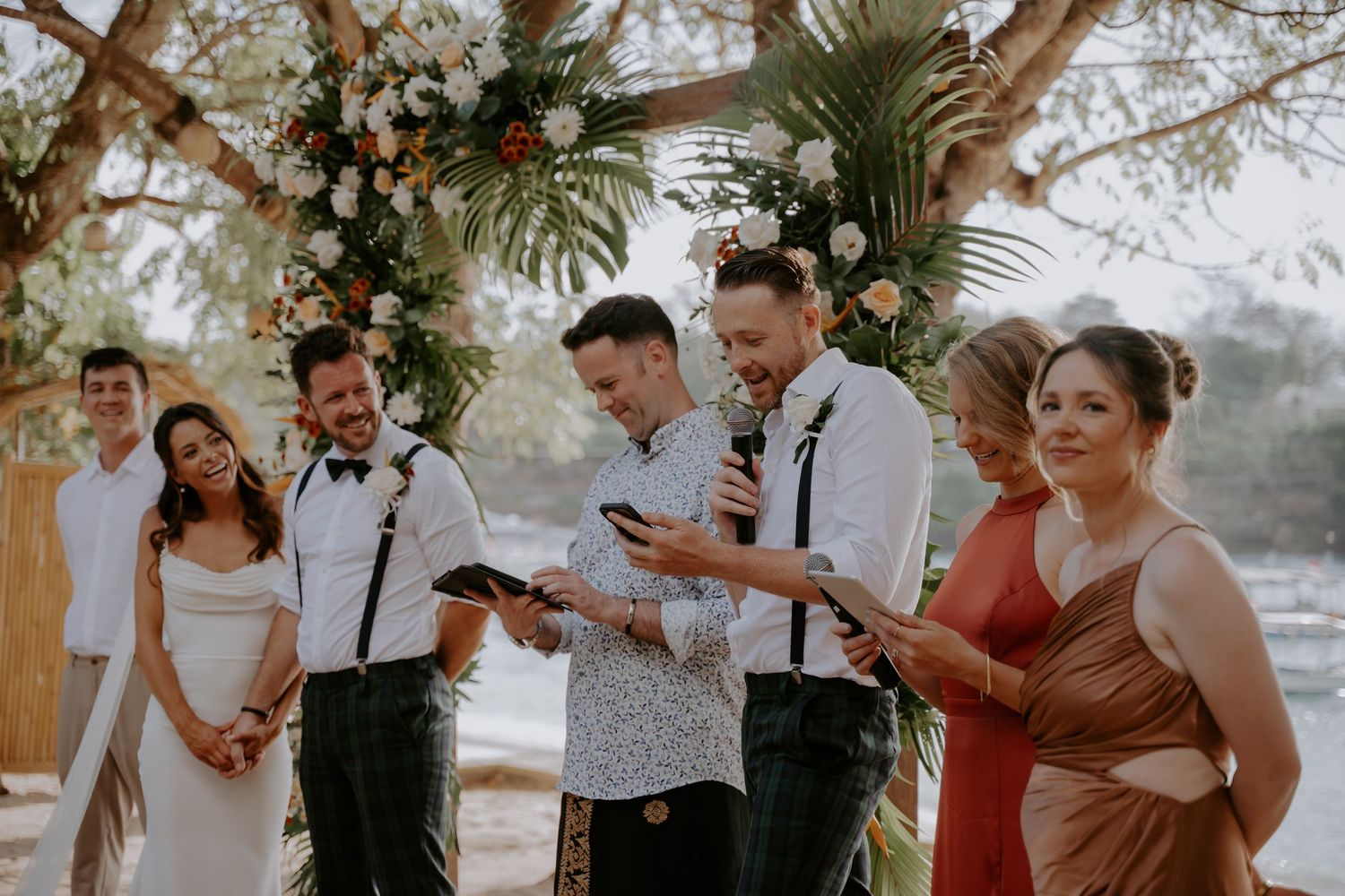 A group celebrates joyfully during a wedding ceremony under a wooden pergola with palm trees and greenery.