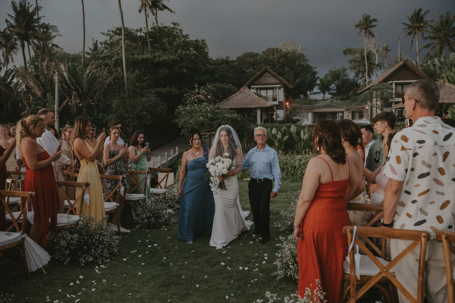 Outdoor wedding ceremony at a tropical venue with palm trees and guests throwing flower petals during celebration.