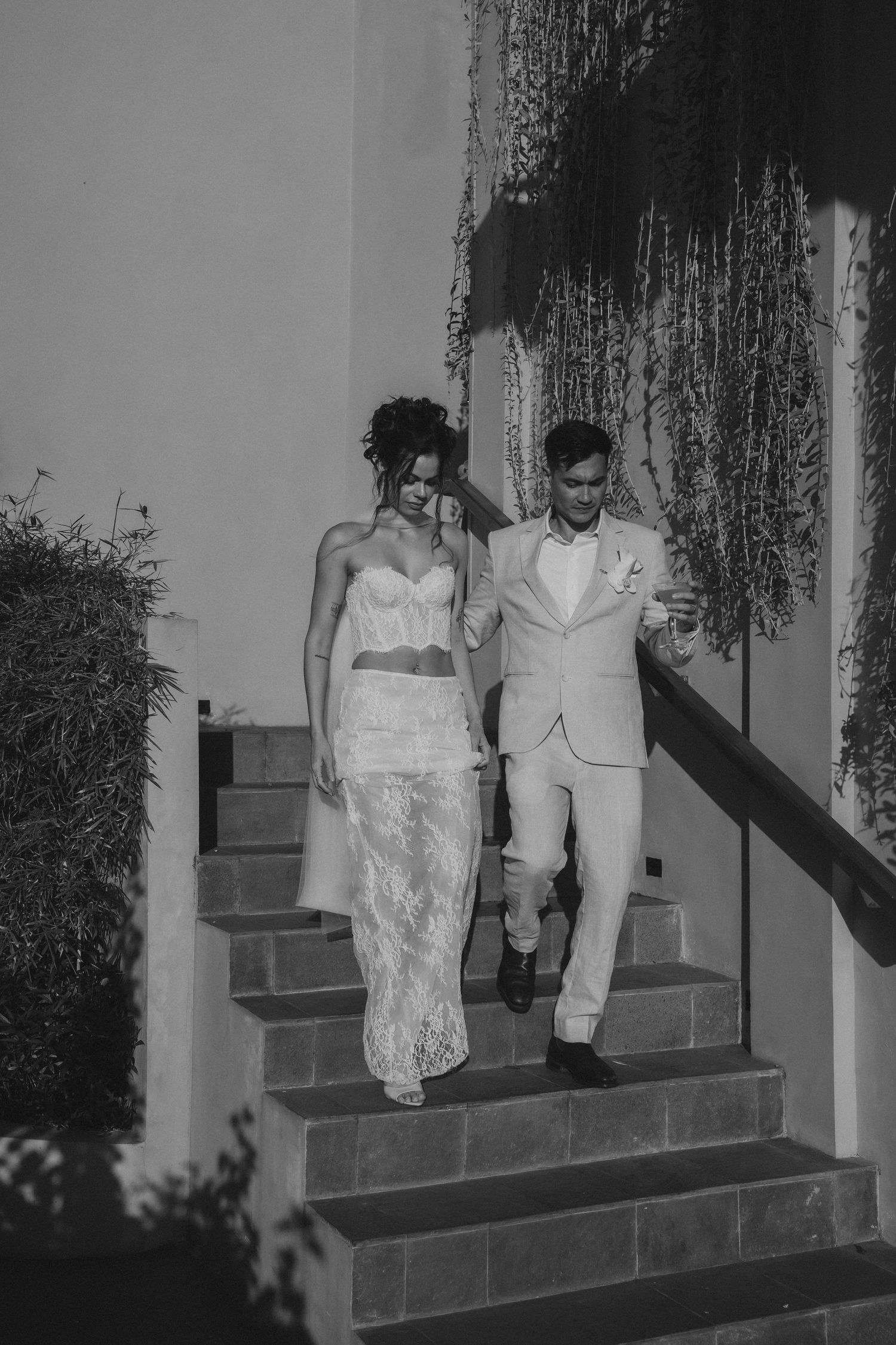 Black and white photo of a couple descending outdoor stairs, with bride wearing lace dress and groom in light suit.