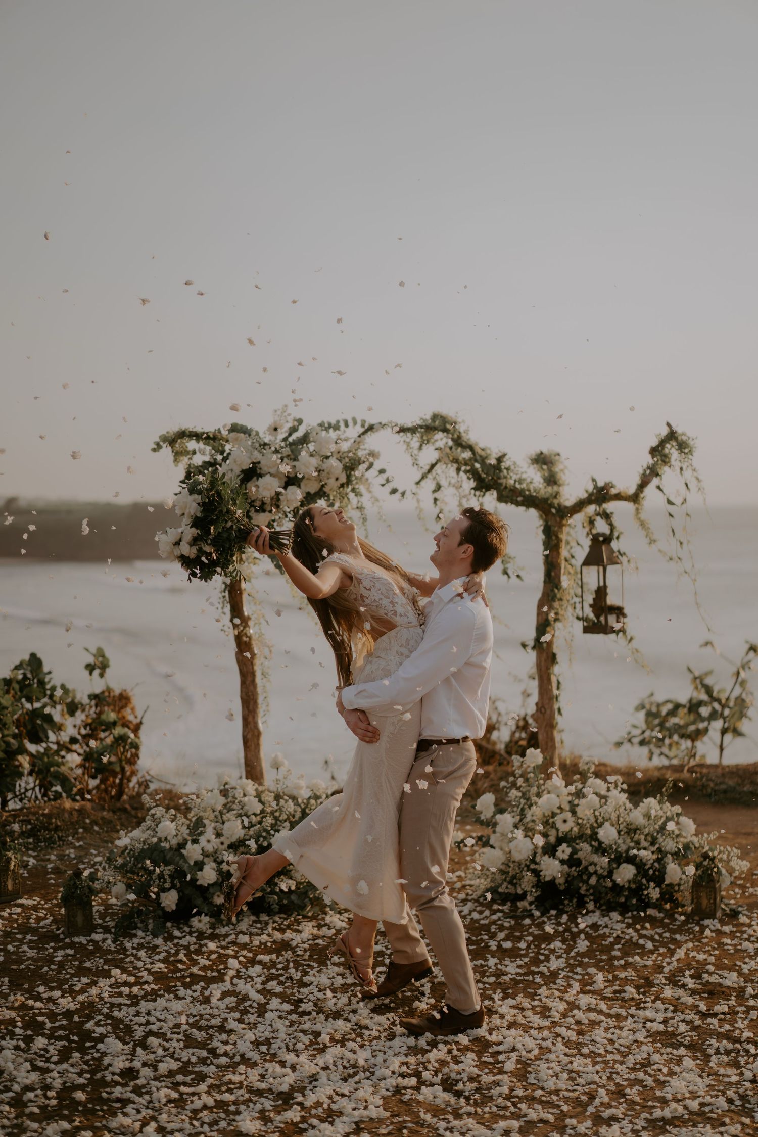 A couple embraces in a romantic moment at sunset under a floral arch by the ocean.