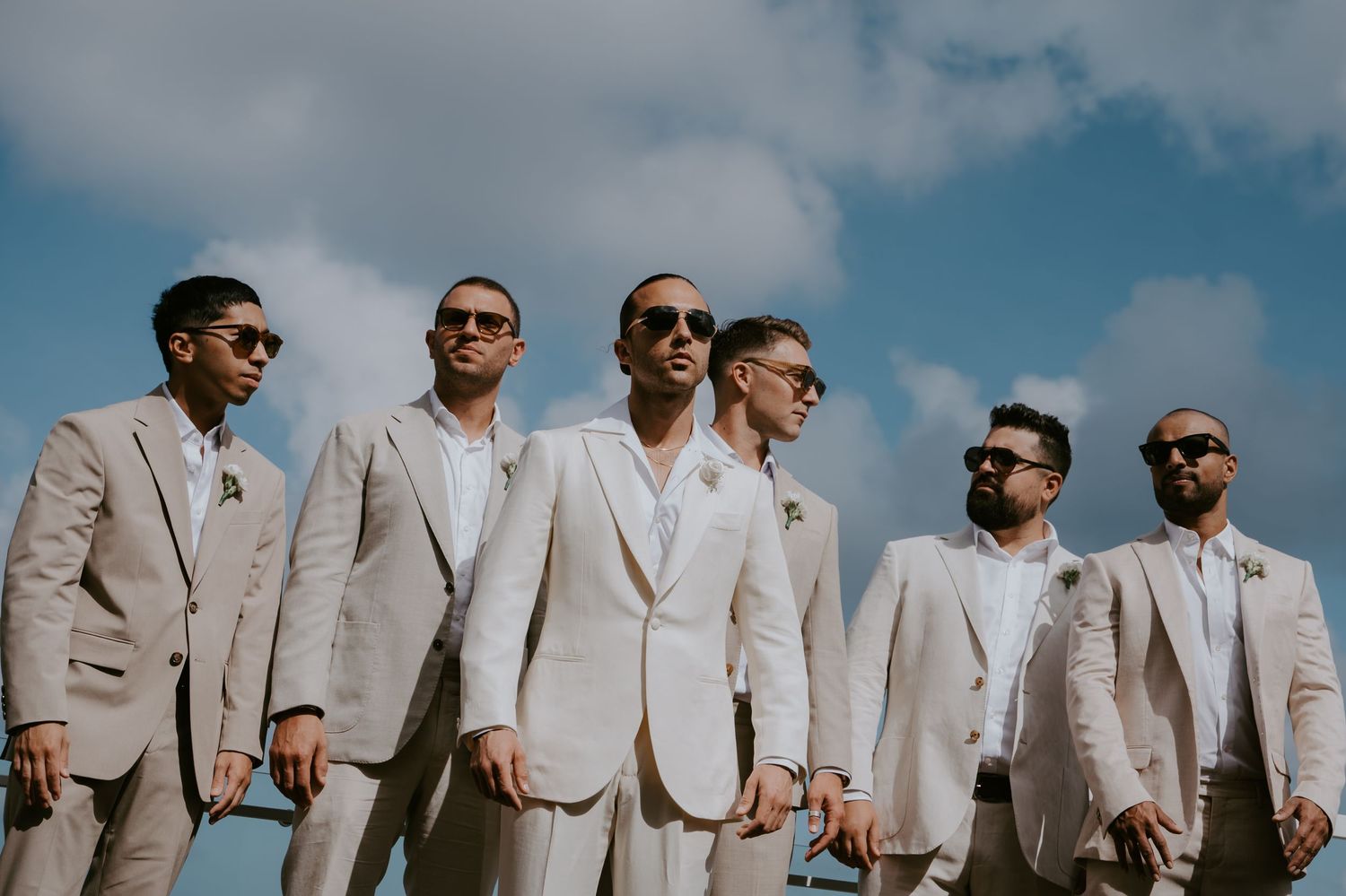 Groomsmen in light beige suits and sunglasses pose together outdoors against a blue cloudy sky.