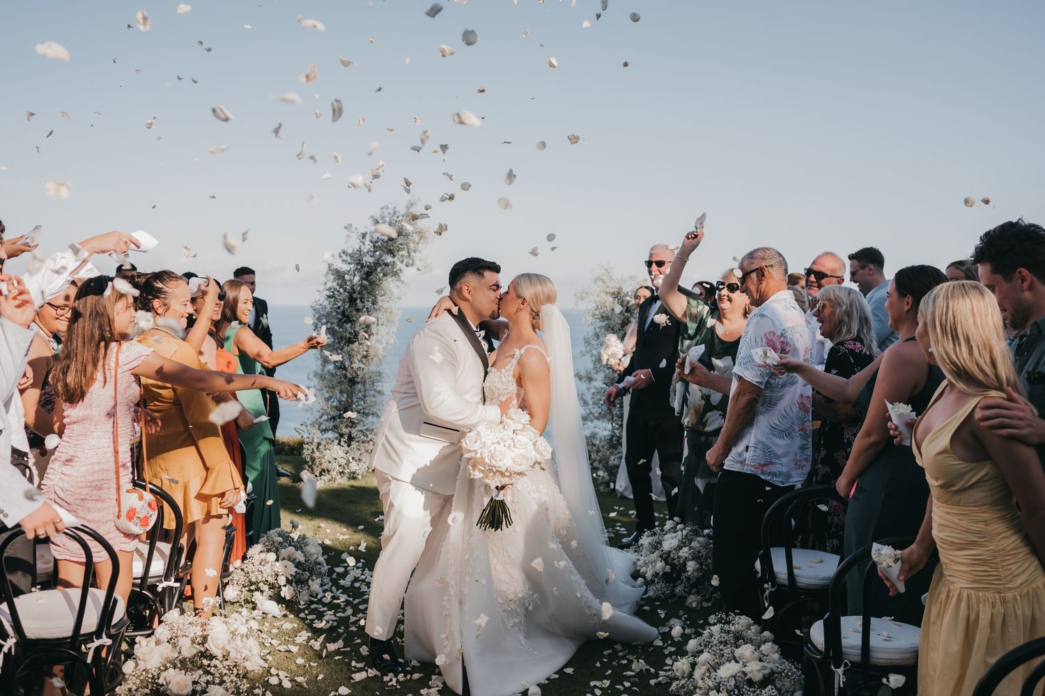 Wedding guests celebrate with confetti toss during outdoor ceremony as newlyweds walk down the aisle.