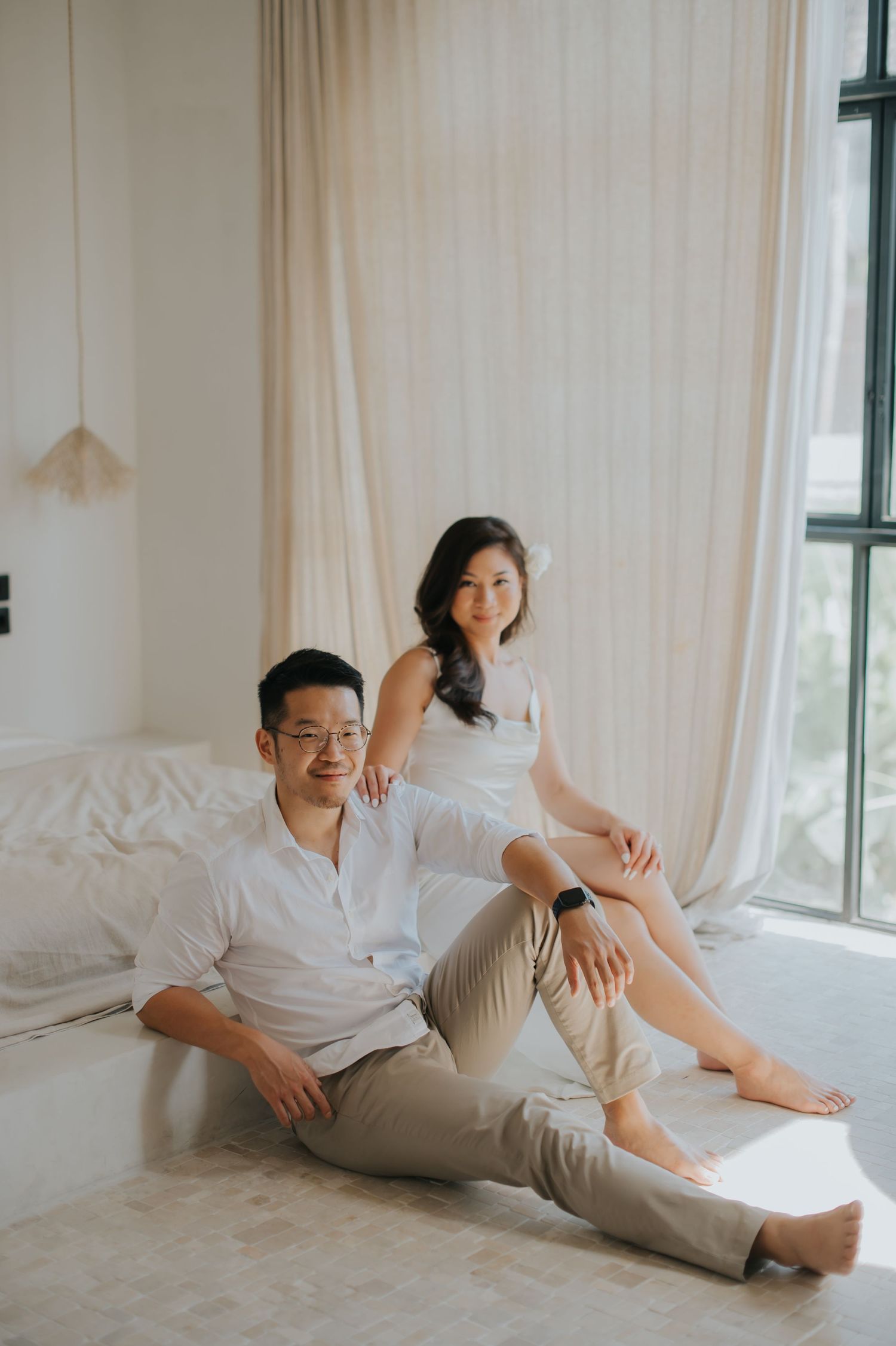A couple in white outfits poses in a bright bedroom, man sitting on floor, woman perched on bed edge.
